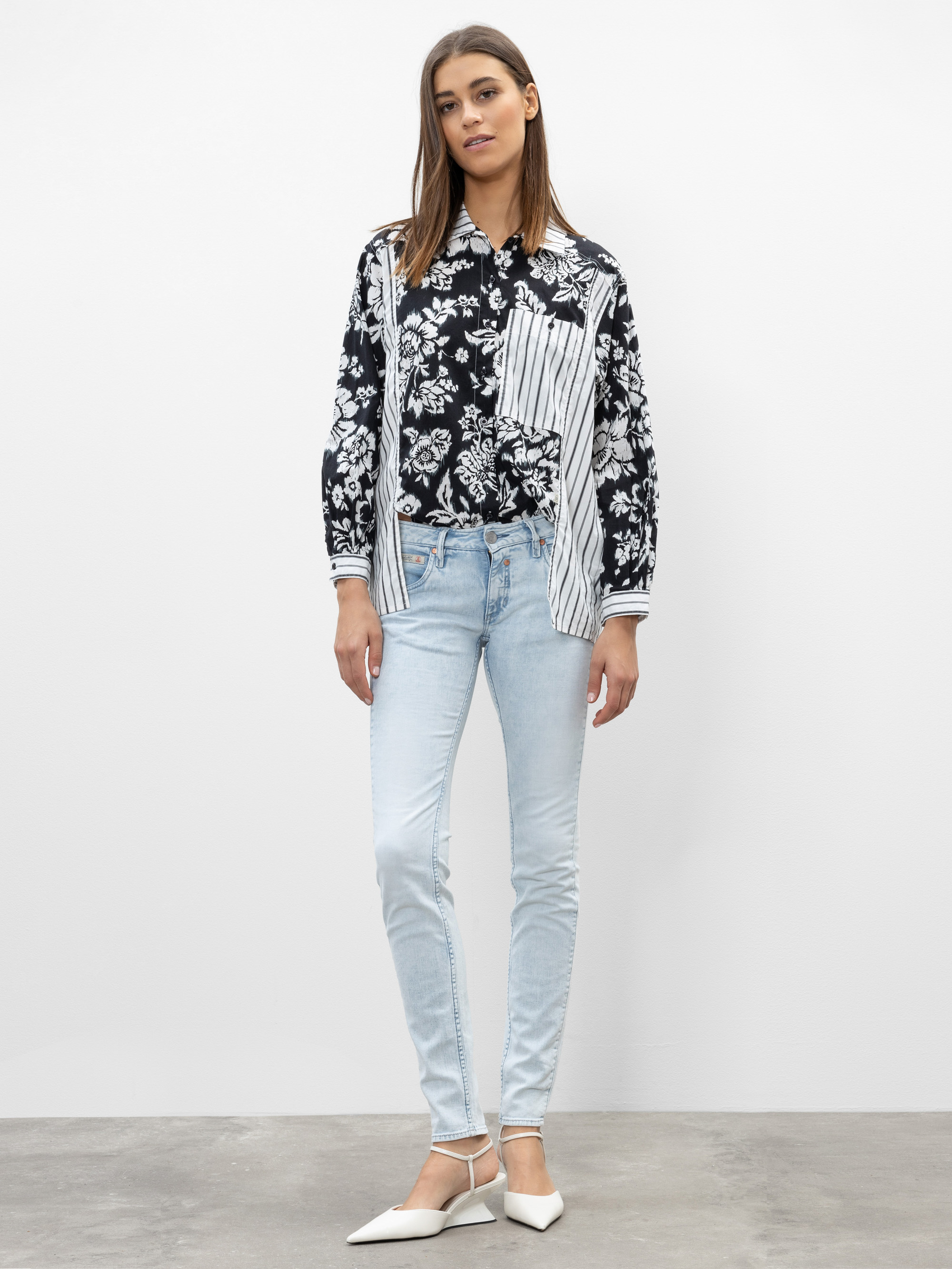 Eine Frau steht vor einer schlichten weißen Wand. Sie trägt ein schwarz-weiß geblümtes und gestreiftes Hemd, hellblaue Jeans und weiße Schuhe mit spitzen Zehen. Ihre Arme liegen entspannt an ihren Seiten, und sie hat glattes braunes Haar.