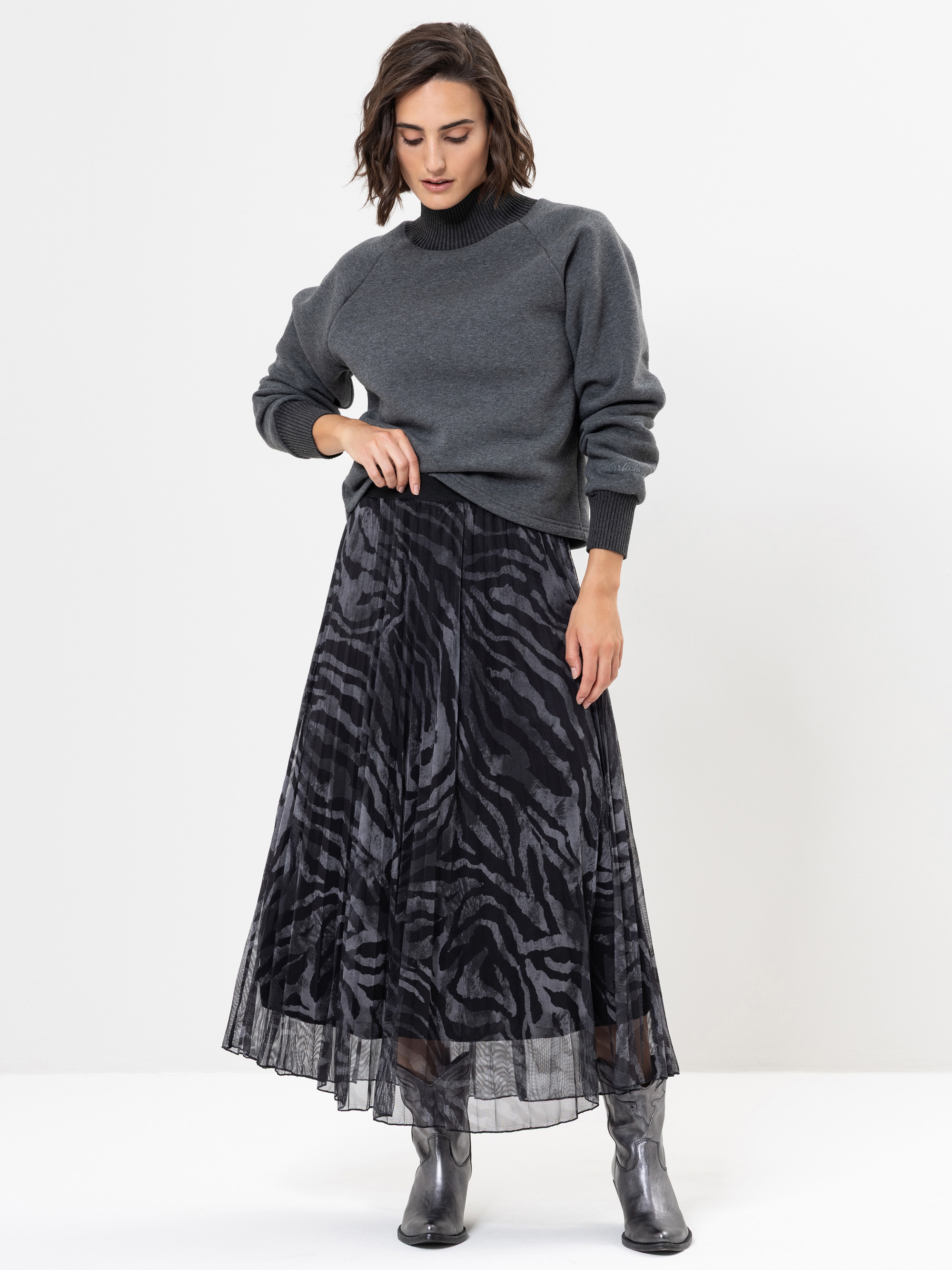 Eine Frau, die einen grauen Pullover und einen langen schwarzen Rock mit Zebramuster trägt, steht vor einem schlichten weißen Hintergrund. Sie kombiniert das Outfit mit glänzenden grauen Stiefeln und blickt mit einer Hand auf ihren Rock herab.