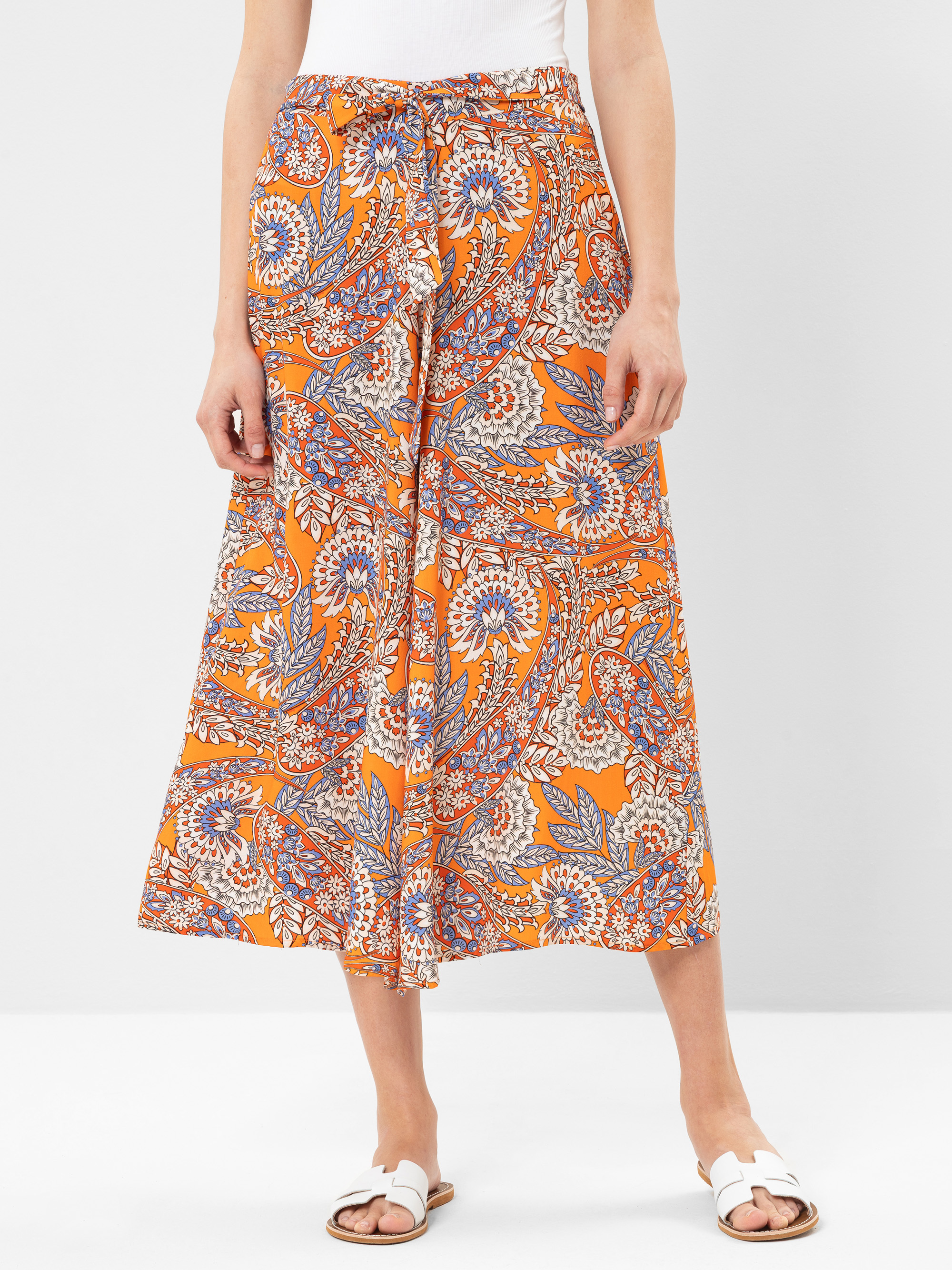 Eine Person, die eine orangefarbene, gemusterte Hose mit weitem Bein und hoher Taille mit Paisley- und Blumenmuster, ein weißes Oberteil und weiße Sandalen trägt, steht vor einem schlichten hellen Hintergrund.