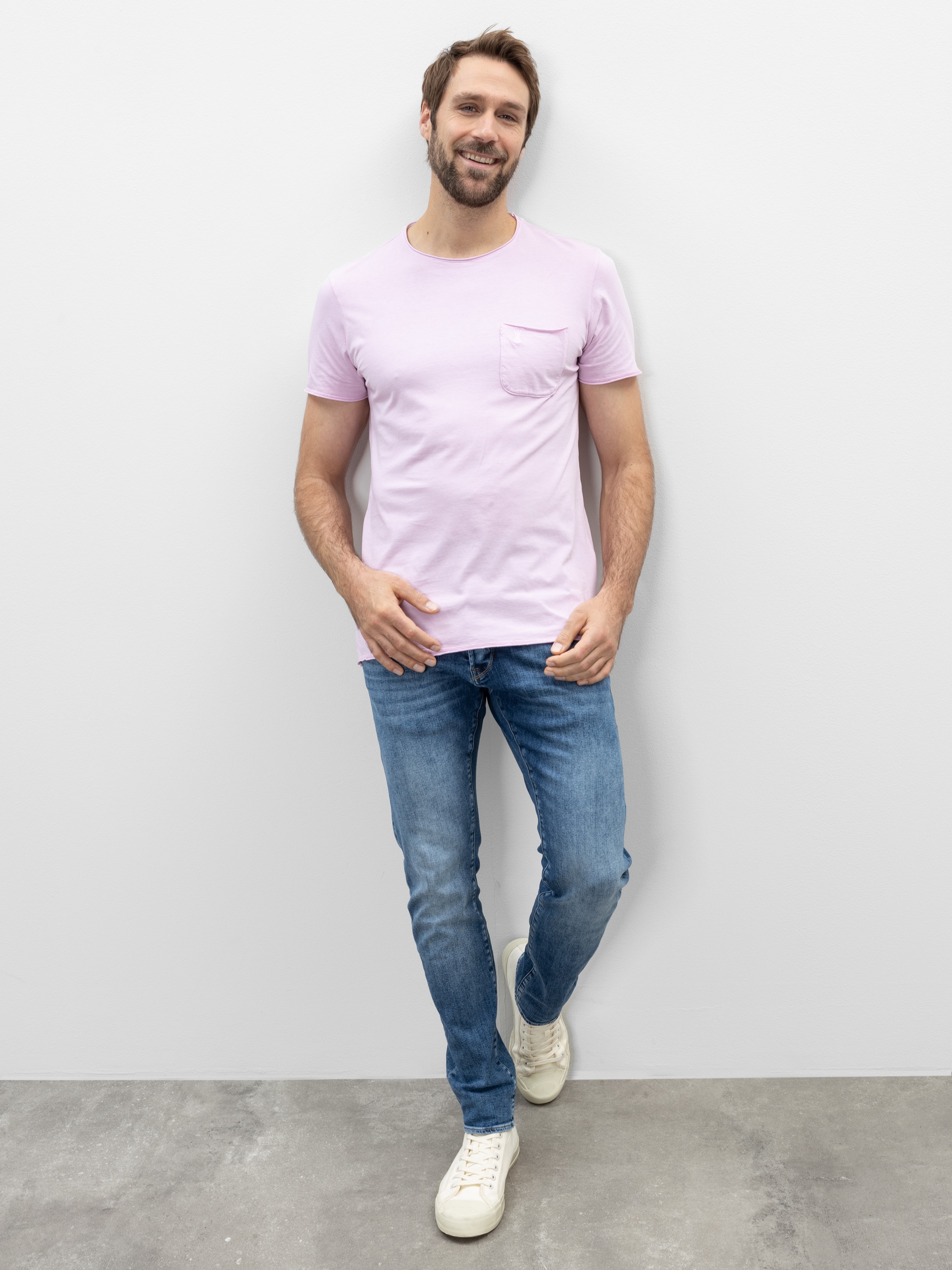 Ein Mann mit kurzen braunen Haaren und einem Bart lächelt, er trägt ein hellrosa T-Shirt, blaue Jeans und weiße Turnschuhe. Er steht an einer schlichten hellen Wand und hat ein Bein leicht vor dem anderen gekreuzt.