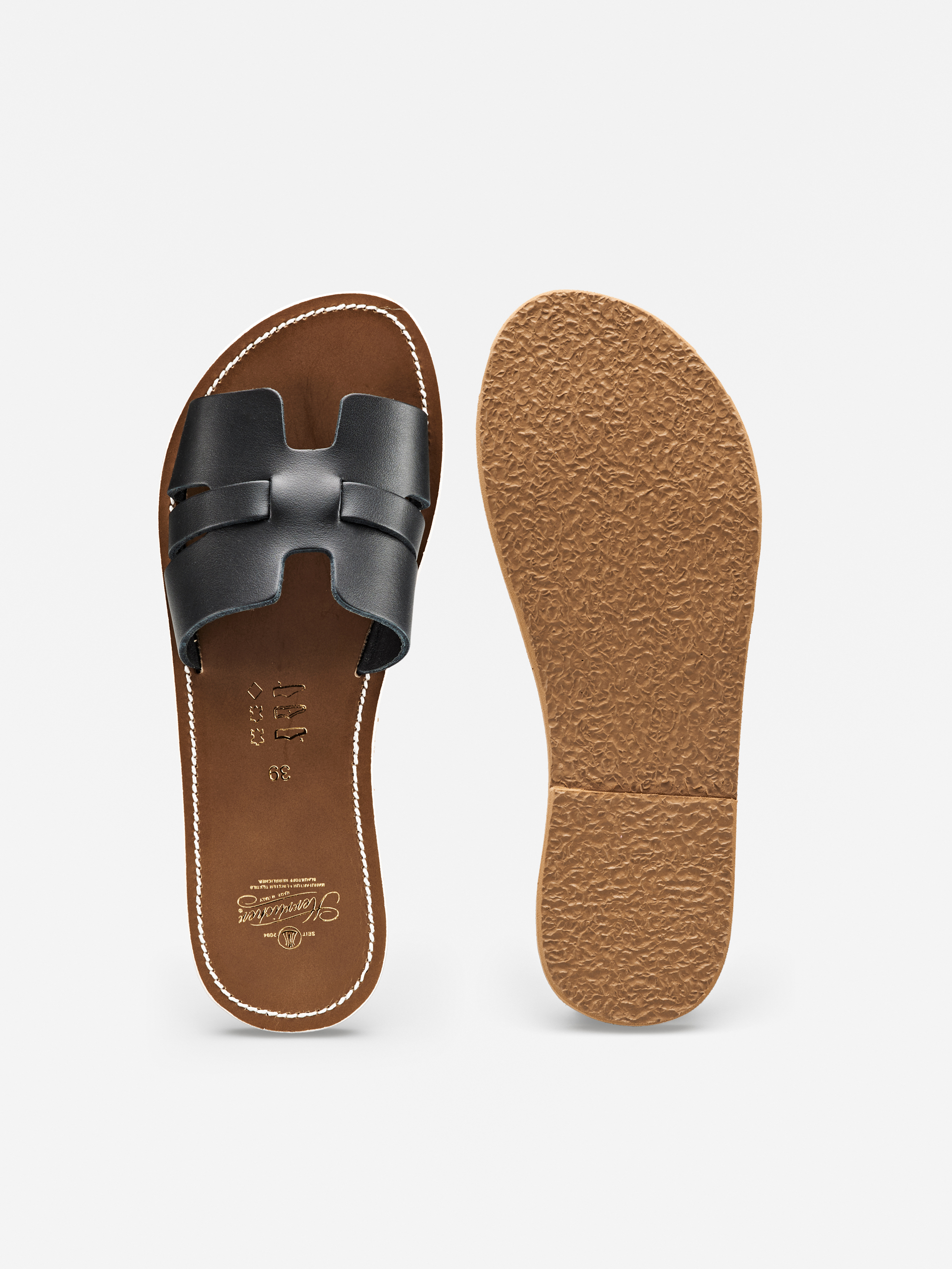 Ein Paar flache Schlupfsandalen mit brauner Sohle und schwarzen, H-förmigen Lederriemen; eine Sandale ist von oben und die andere von unten gezeigt, wobei die strukturierte Sohle zu sehen ist.
