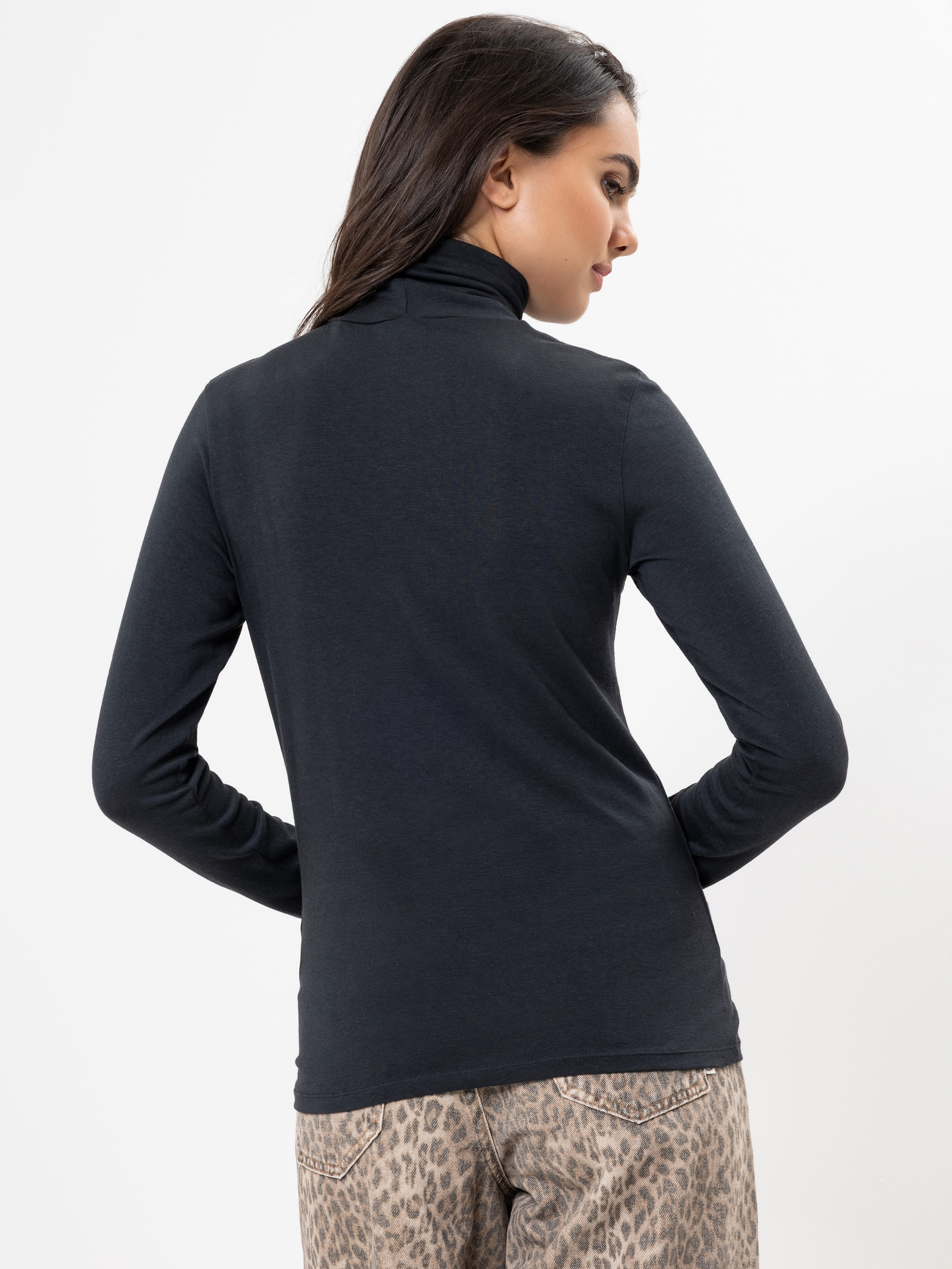 Eine Frau mit langen dunklen Haaren trägt ein schwarzes langärmeliges Rollkragenoberteil und eine helle Hose mit Leopardenmuster. Sie blickt von der Kamera weg auf einen schlichten weißen Hintergrund.