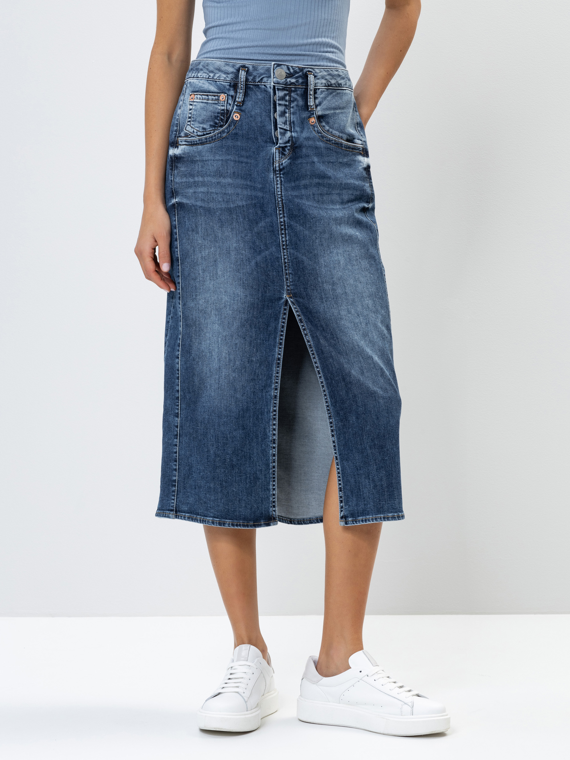 Eine Person, die ein hellblaues, geripptes Oberteil, einen hochgeschlossenen, dunklen Jeans-Midirock mit einem Schlitz an der Vorderseite und weiße Turnschuhe trägt, steht vor einem schlichten weißen Hintergrund.