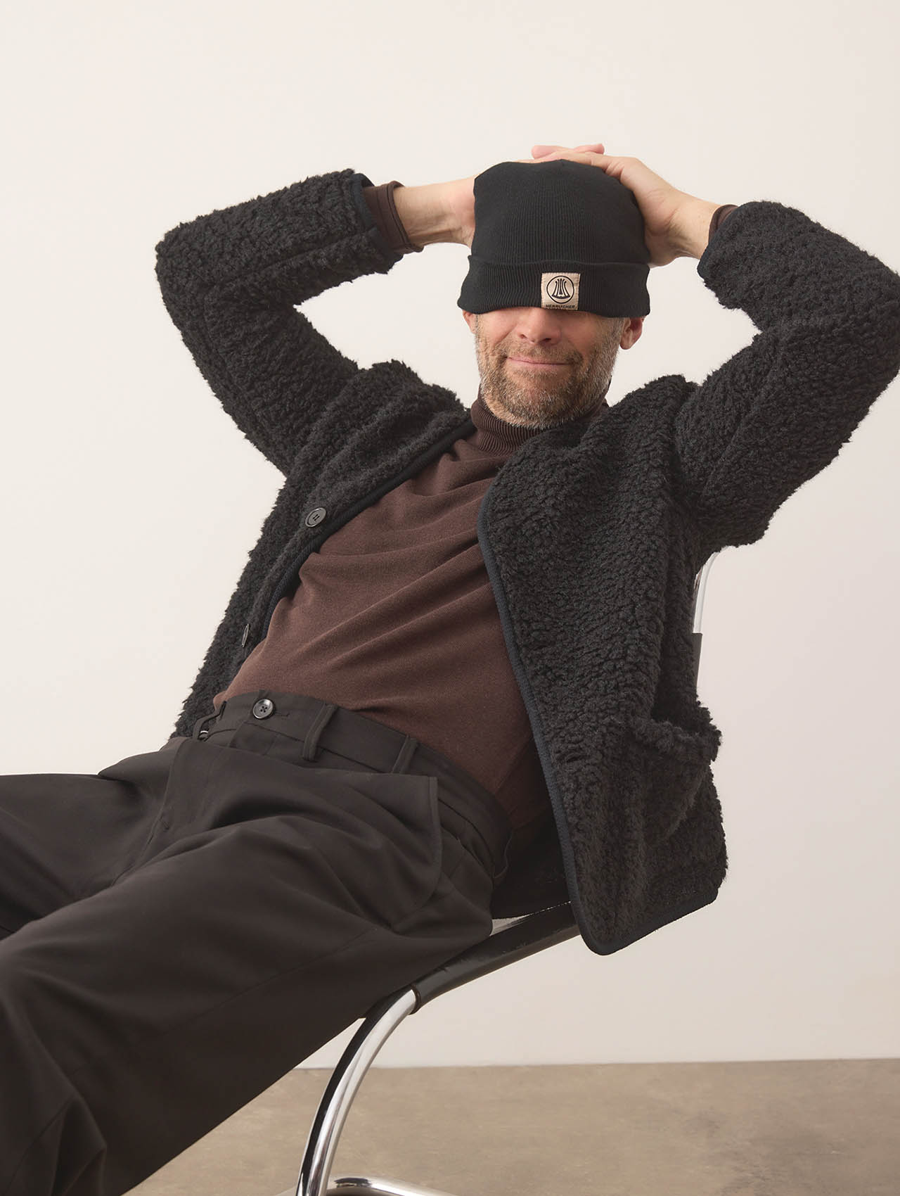 Ein Mann sitzt auf einem Stuhl, lehnt sich zurück und lächelt, während er sich mit den Händen eine schwarze Mütze über die Augen stülpt. Er trägt eine strukturierte schwarze Jacke, ein braunes Hemd und eine schwarze Hose vor einem schlichten Hinte