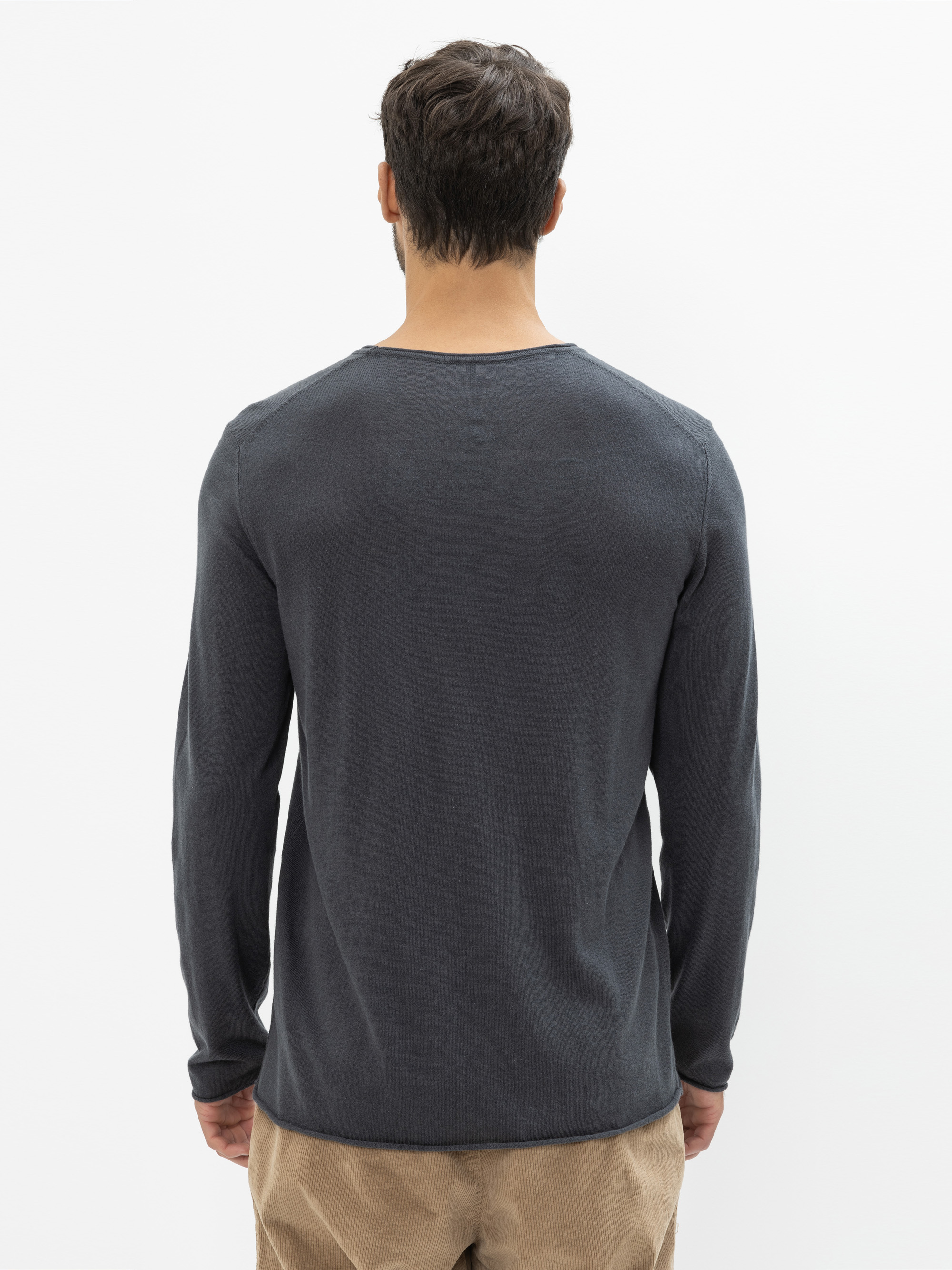 Ein Mann mit kurzen braunen Haaren steht mit dem Rücken zur Kamera, trägt ein langärmeliges dunkelgraues Hemd und eine hellbraune Hose vor einem schlichten weißen Hintergrund.