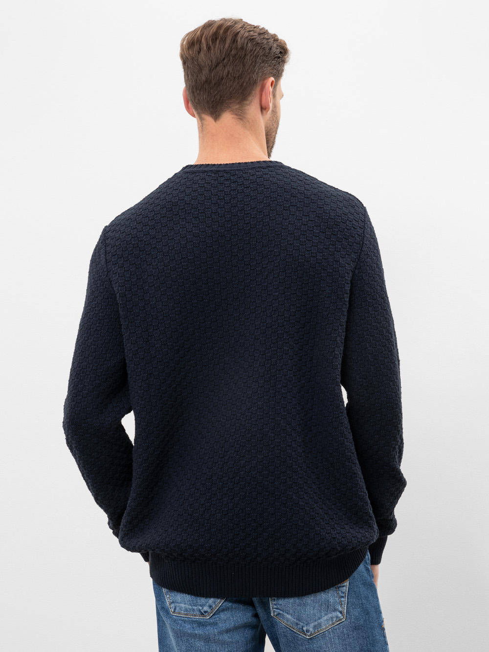 Ein Mann mit kurzen braunen Haaren steht mit dem Rücken zur Kamera. Er trägt einen strukturierten dunkelnavyfarbenen Pullover und blaue Jeans vor einem schlichten weißen Hintergrund.