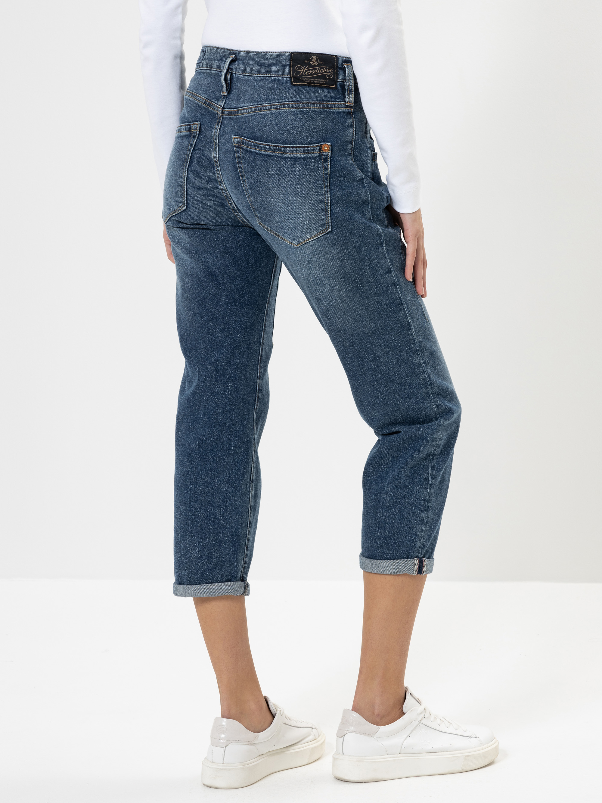 Person, die eine blaue Jeans mit Stulpen, ein weißes langärmeliges Oberteil und weiße Turnschuhe trägt, stehend vor einem schlichten weißen Hintergrund, von hinten gesehen.