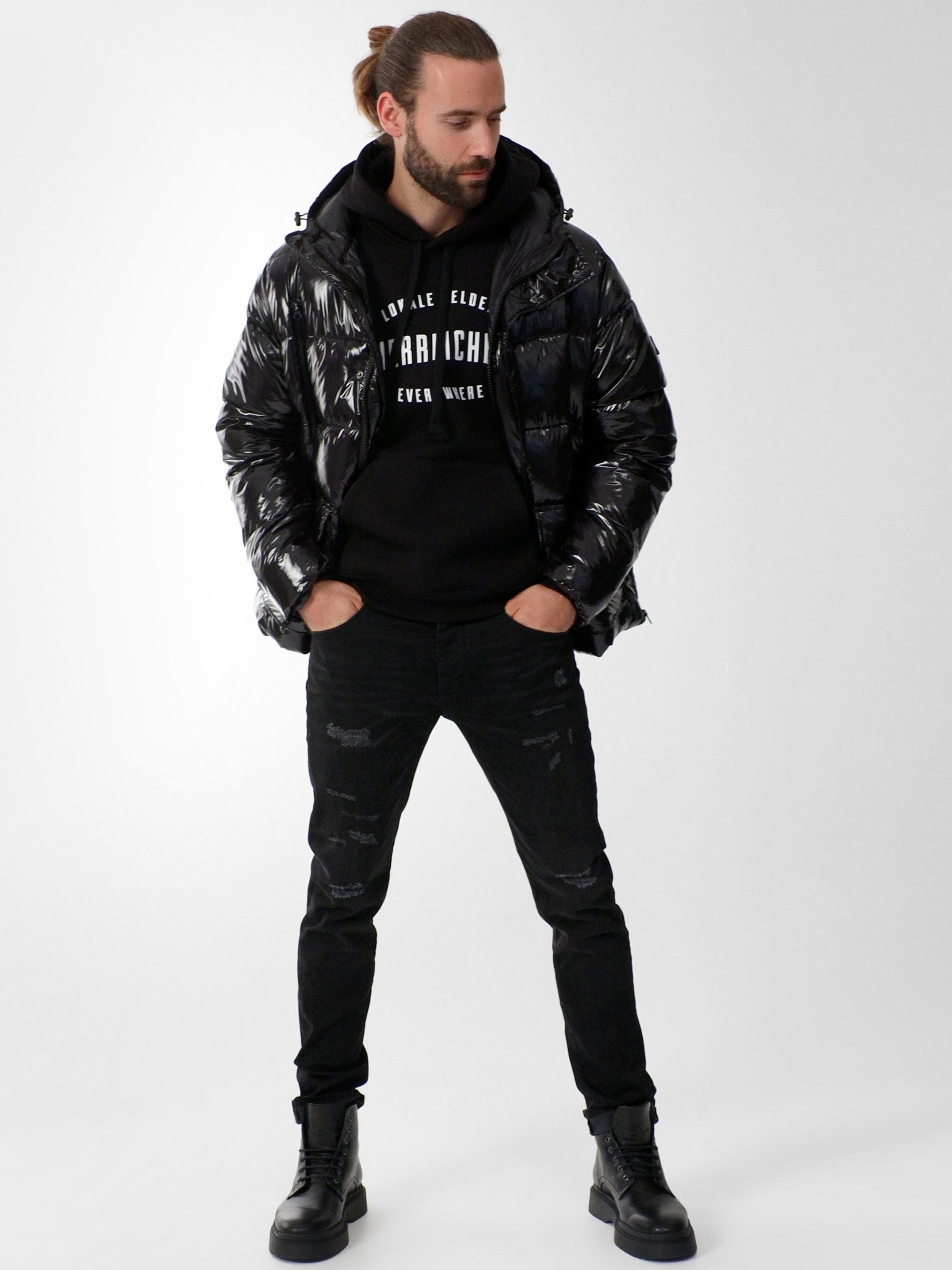 Ein Mann mit Bart und zurückgebundenem Haar trägt eine glänzende schwarze Pufferjacke über einem schwarzen Kapuzenpulli, schwarze Jeans und schwarze Stiefel und steht vor einem schlichten hellen Hintergrund.