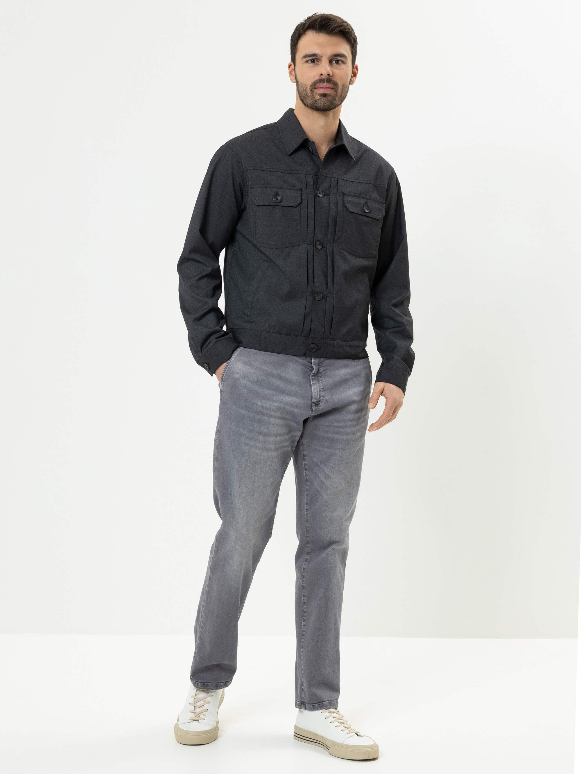 Ein Mann mit kurzen dunklen Haaren und einem Bart steht vor einem weißen Hintergrund und trägt ein schwarzes Button-up-Hemd, hellgraue Jeans und weiße Turnschuhe. Er hat eine Hand in der Tasche und schaut in die Kamera.