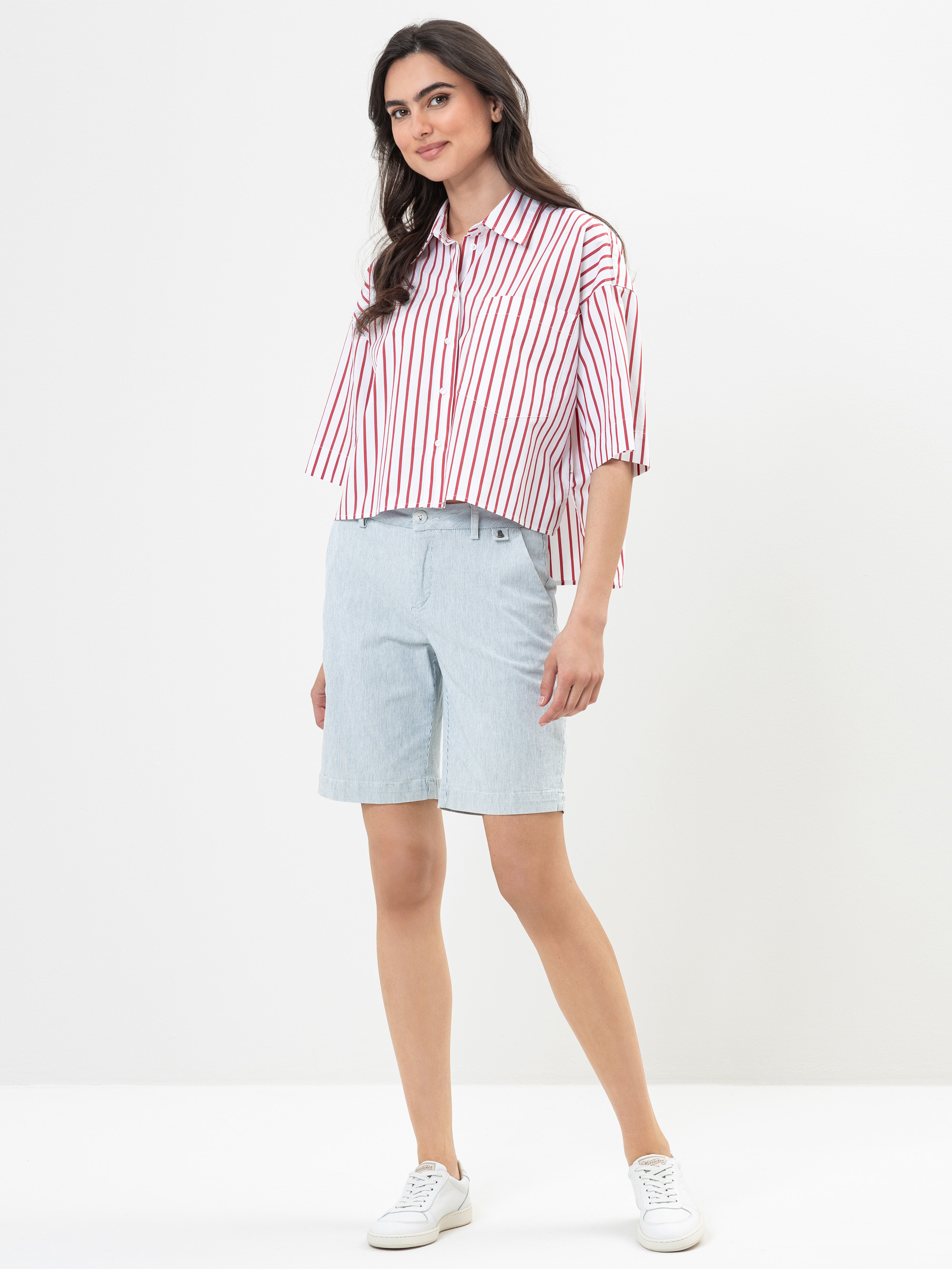 Eine Frau steht vor einem schlichten weißen Hintergrund und trägt ein locker sitzendes, rot-weiß senkrecht gestreiftes Hemd, hellblaue knielange Shorts und weiße Turnschuhe. Sie lächelt und hat langes, dunkles Haar.