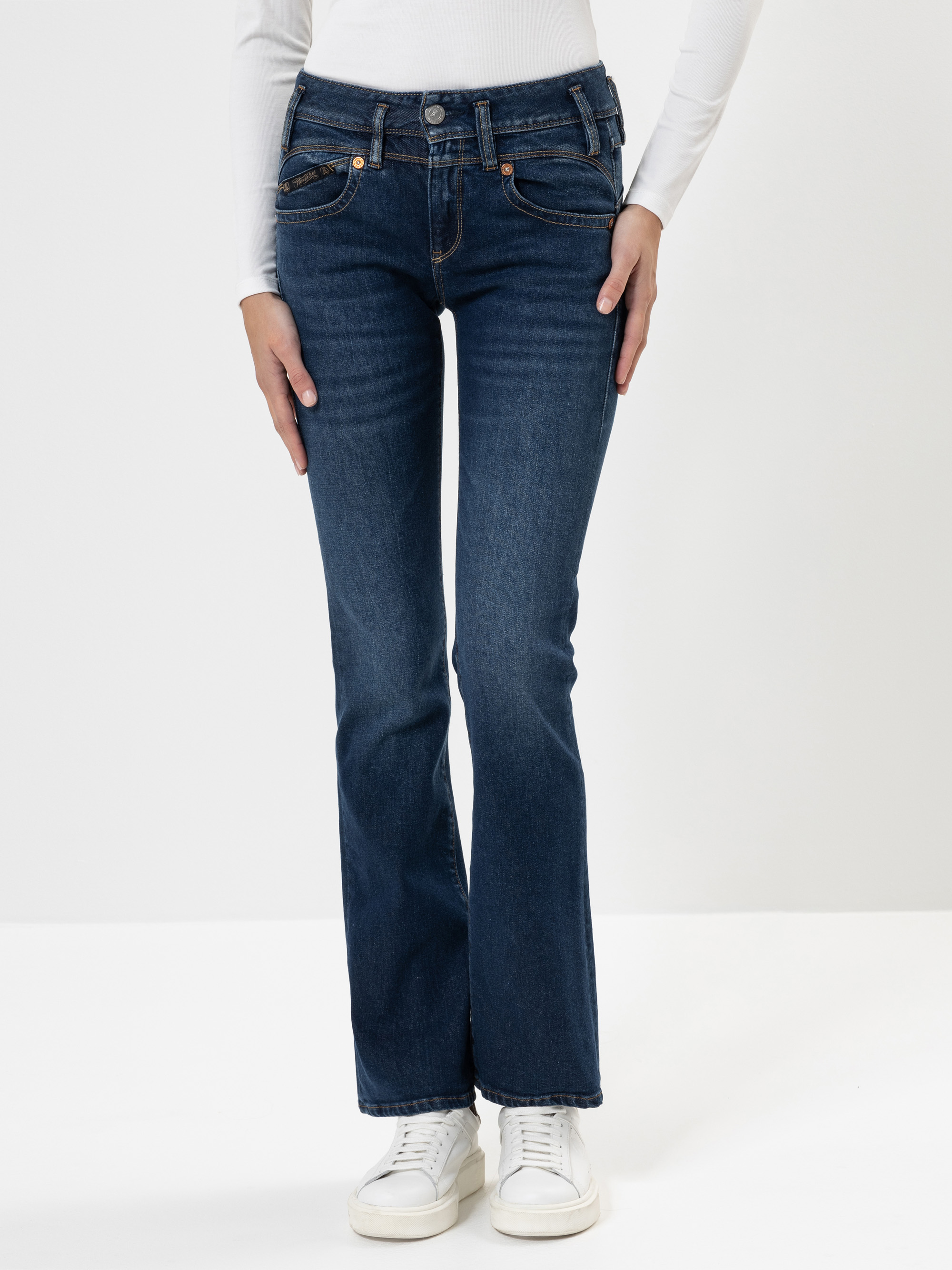 Eine Person, die dunkelblaue Bootcut-Jeans, ein weißes langärmeliges Oberteil und weiße Turnschuhe trägt, steht vor einem schlichten weißen Hintergrund und hat die Hände leicht an die Seite gelegt.