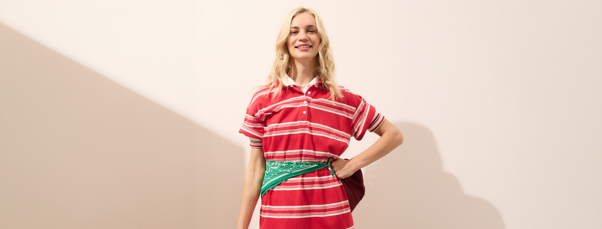 Eine lächelnde Person mit langen blonden Haaren trägt ein rot-weiß gestreiftes Polohemd und ein grünes Kopftuch um die Taille gebunden und steht vor einem schlichten hellen Hintergrund.