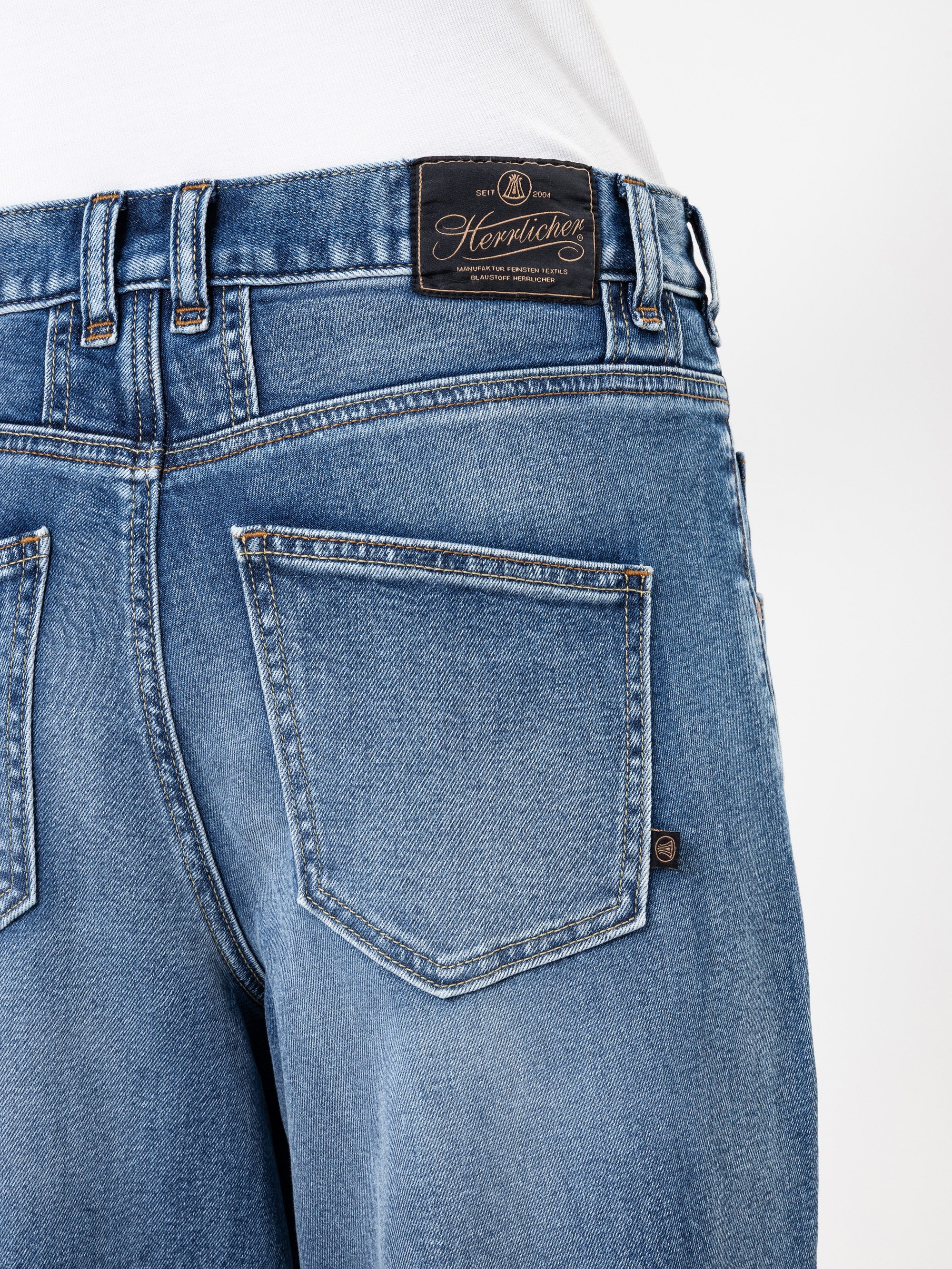 Nahaufnahme des Rückens einer Person, die eine blaue Denim-Jeans trägt, mit zwei Gesäßtaschen und einem schwarzen Lederaufnäher mit dem Markennamen Herrlicher über der rechten Tasche. Die Person trägt außerdem ein weißes Hemd.