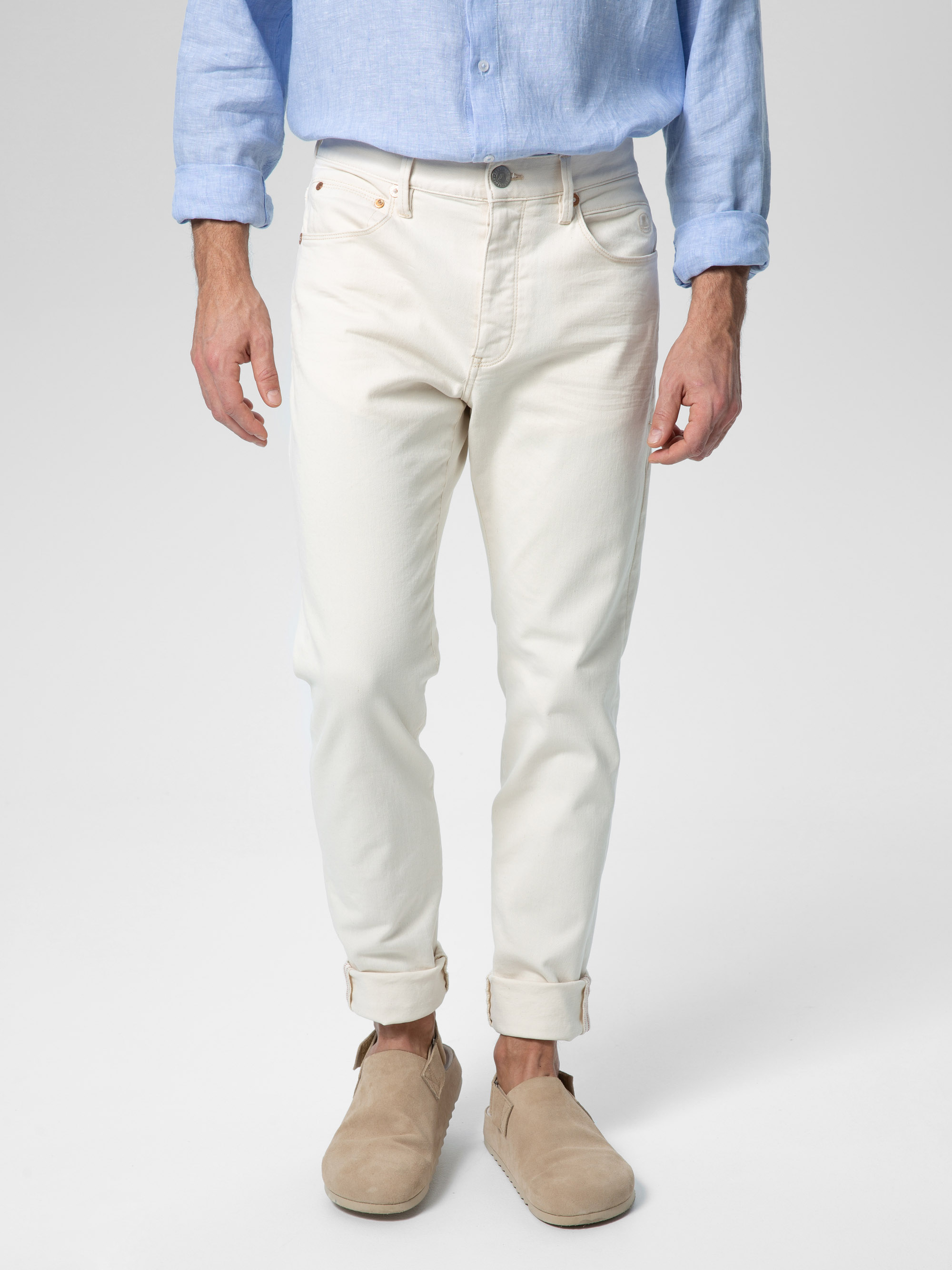 Eine Person, die ein hellblaues Hemd, cremefarbene Jeans mit Bündchen und beigefarbene Slipper trägt, steht vor einem schlichten hellen Hintergrund. Der Kopf der Person ist auf dem Bild nicht zu sehen.
