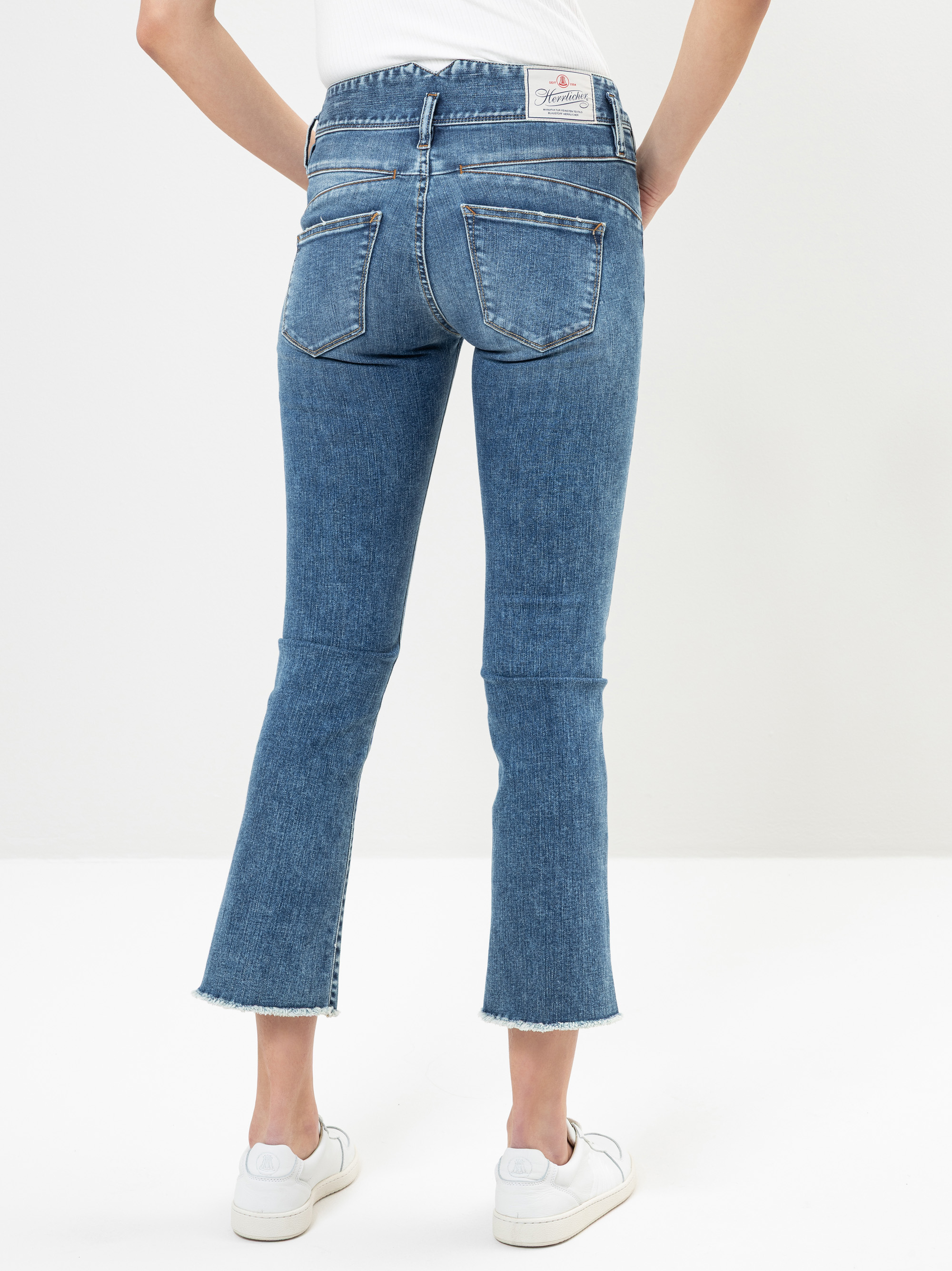 Eine Person in blauen Schlagjeans mit ausgefransten Säumen und weißen Turnschuhen, die vor einem schlichten weißen Hintergrund steht, von hinten gesehen.