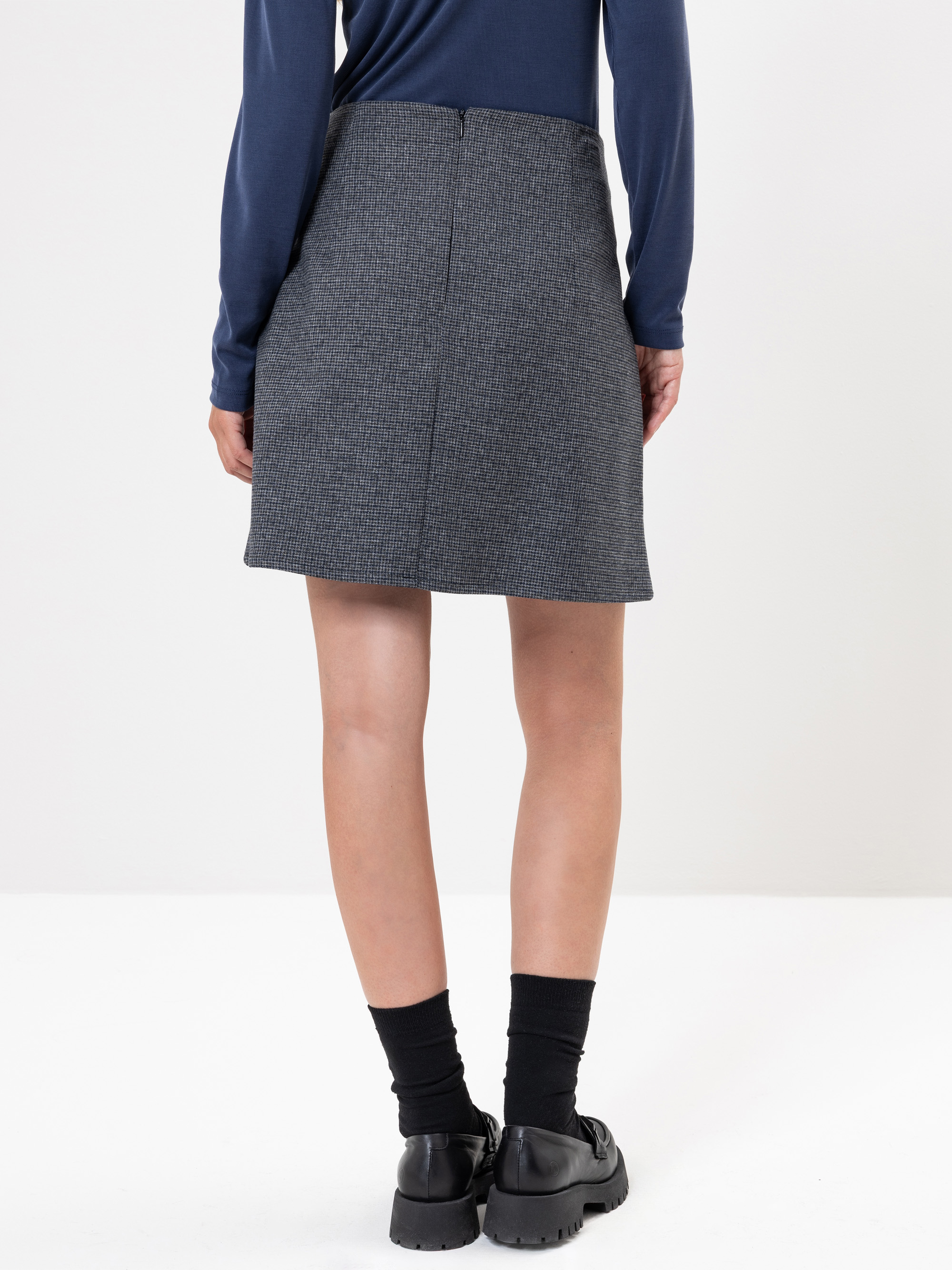 Eine Person steht mit dem Gesicht zur Wand und trägt einen dunkelgrauen Rock, ein marineblaues langärmeliges Oberteil, schwarze Socken und schwarze Schuhe vor einem schlichten weißen Hintergrund.