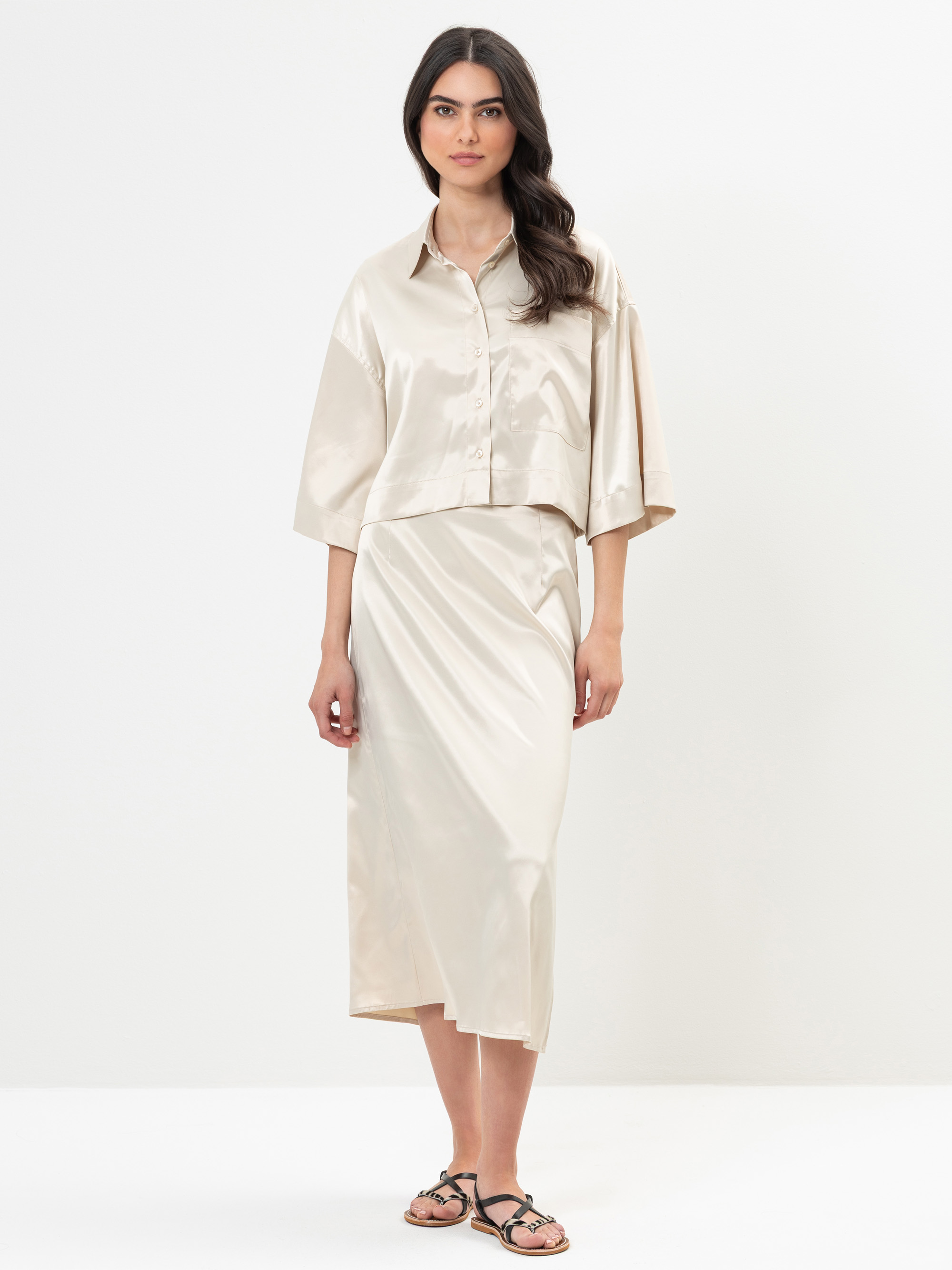 Eine Frau steht vor einem weißen Hintergrund und trägt ein lockeres, hellbeiges Satin-Knopfleistenhemd mit ellenbogenlangen Ärmeln und einen passenden Midirock, dazu schwarze Sandalen. Sie hat langes, dunkles, gewelltes Haar und einen neutralen Gesicht