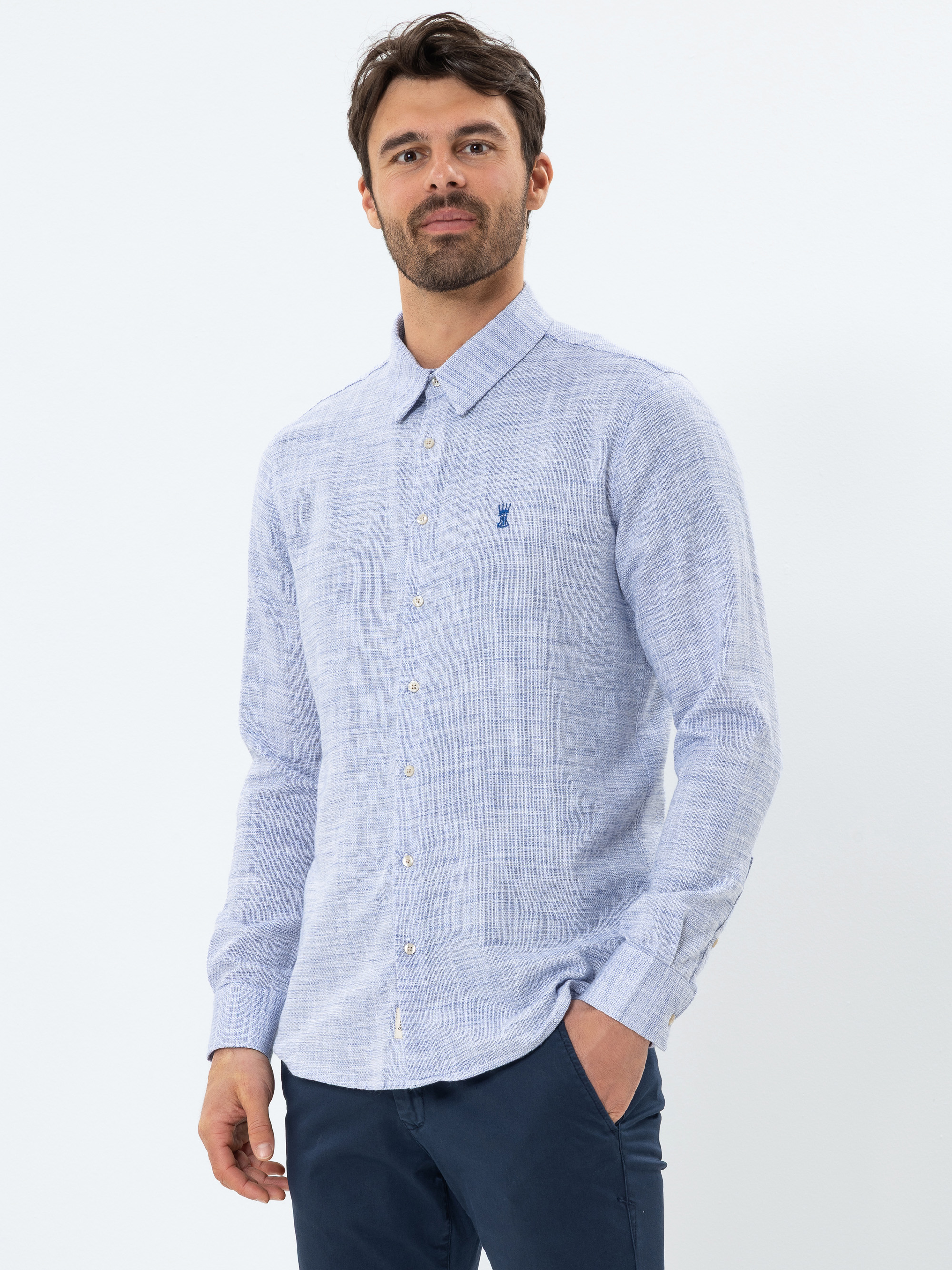 Ein Mann mit dunklem Haar und Bart trägt ein hellblaues, langärmeliges Button-up-Hemd und eine dunkle Hose. Er steht vor einem schlichten weißen Hintergrund und hat eine Hand in der Tasche.