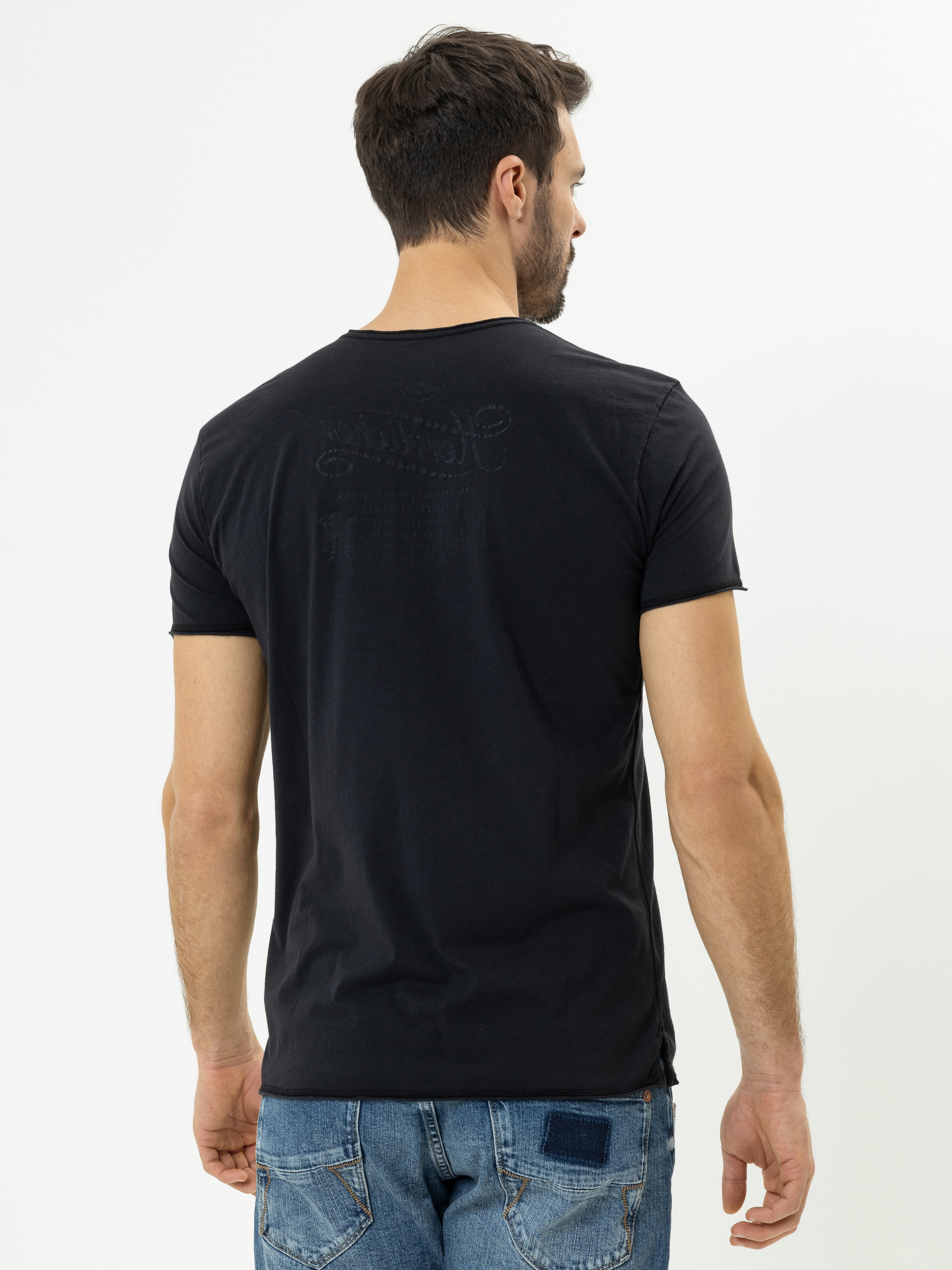 Ein Mann mit kurzen dunklen Haaren, der ein schwarzes T-Shirt und blaue Jeans trägt, steht vor einem schlichten weißen Hintergrund und blickt von der Kamera weg.