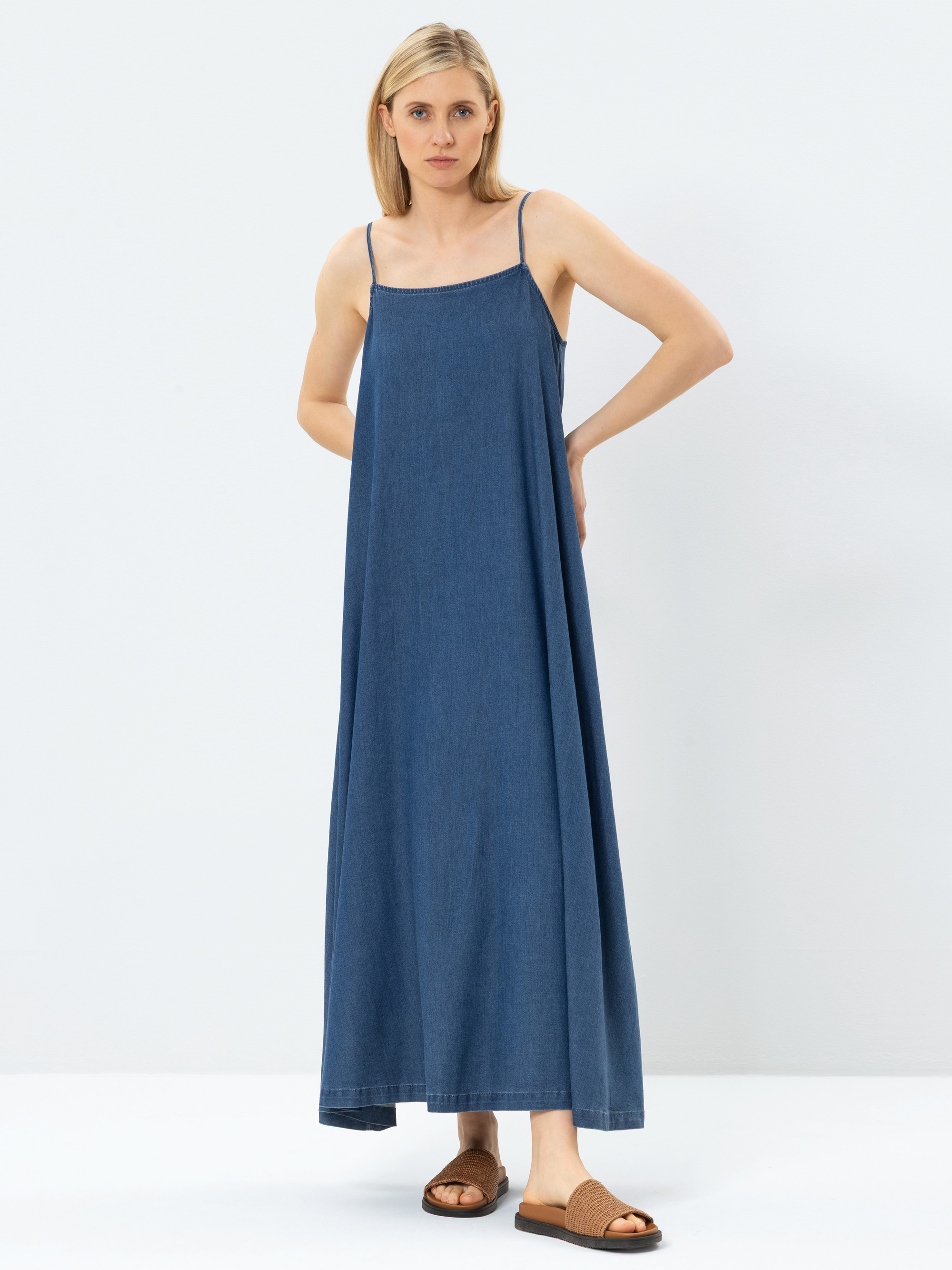 Eine Frau in einem weiten, ärmellosen, blauen Jeans-Maxikleid mit dünnen Trägern steht vor einem schlichten weißen Hintergrund. Sie hat glattes blondes Haar und trägt braune, zehenoffene Slide-Sandalen.
