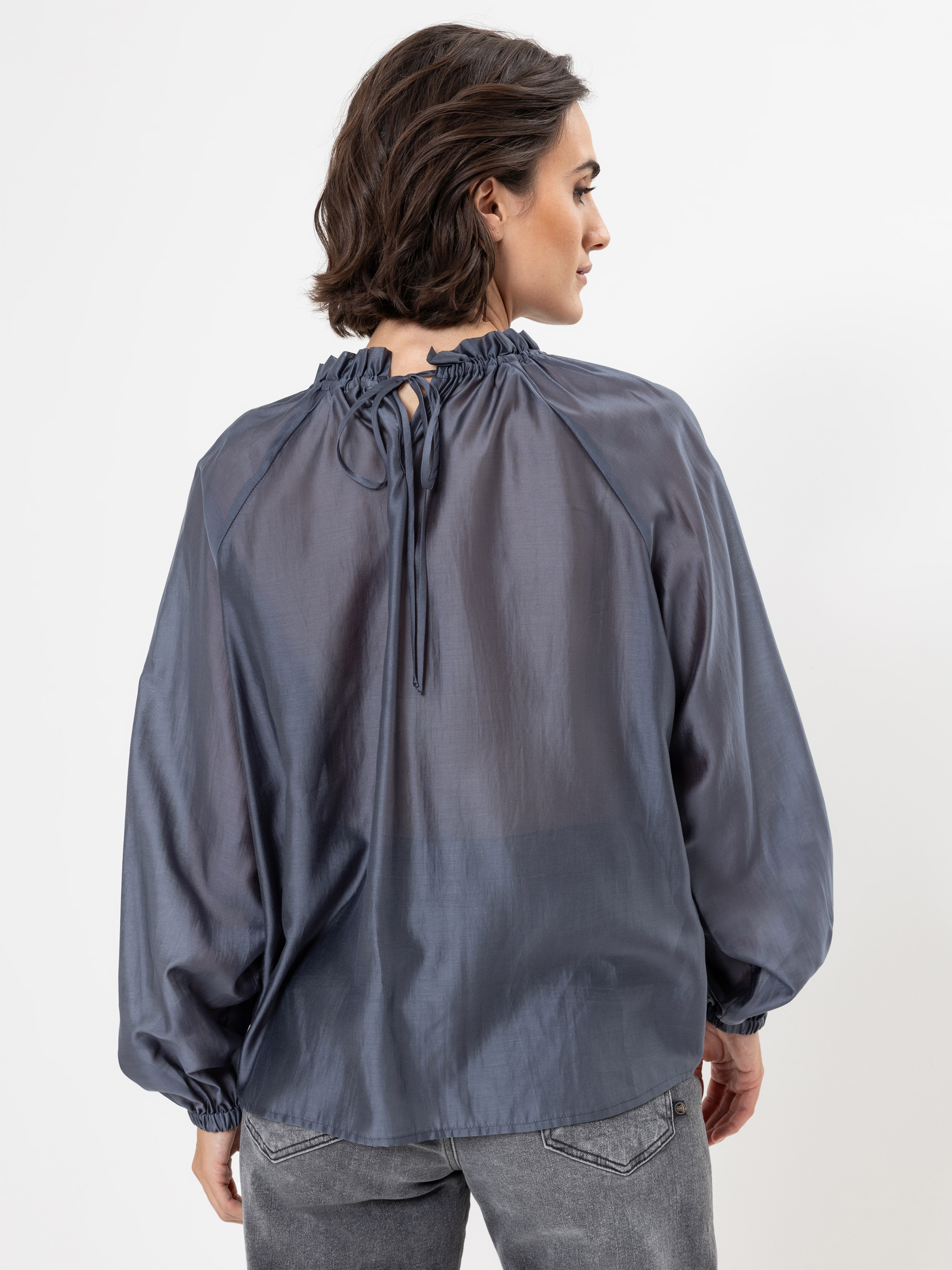 Eine Frau mit kurzen braunen Haaren trägt eine lockere, glänzende, langärmelige blaue Bluse mit gerafftem Ausschnitt und einer Krawatte auf dem Rücken. Sie blickt von der Kamera weg und trägt außerdem eine graue Jeans.