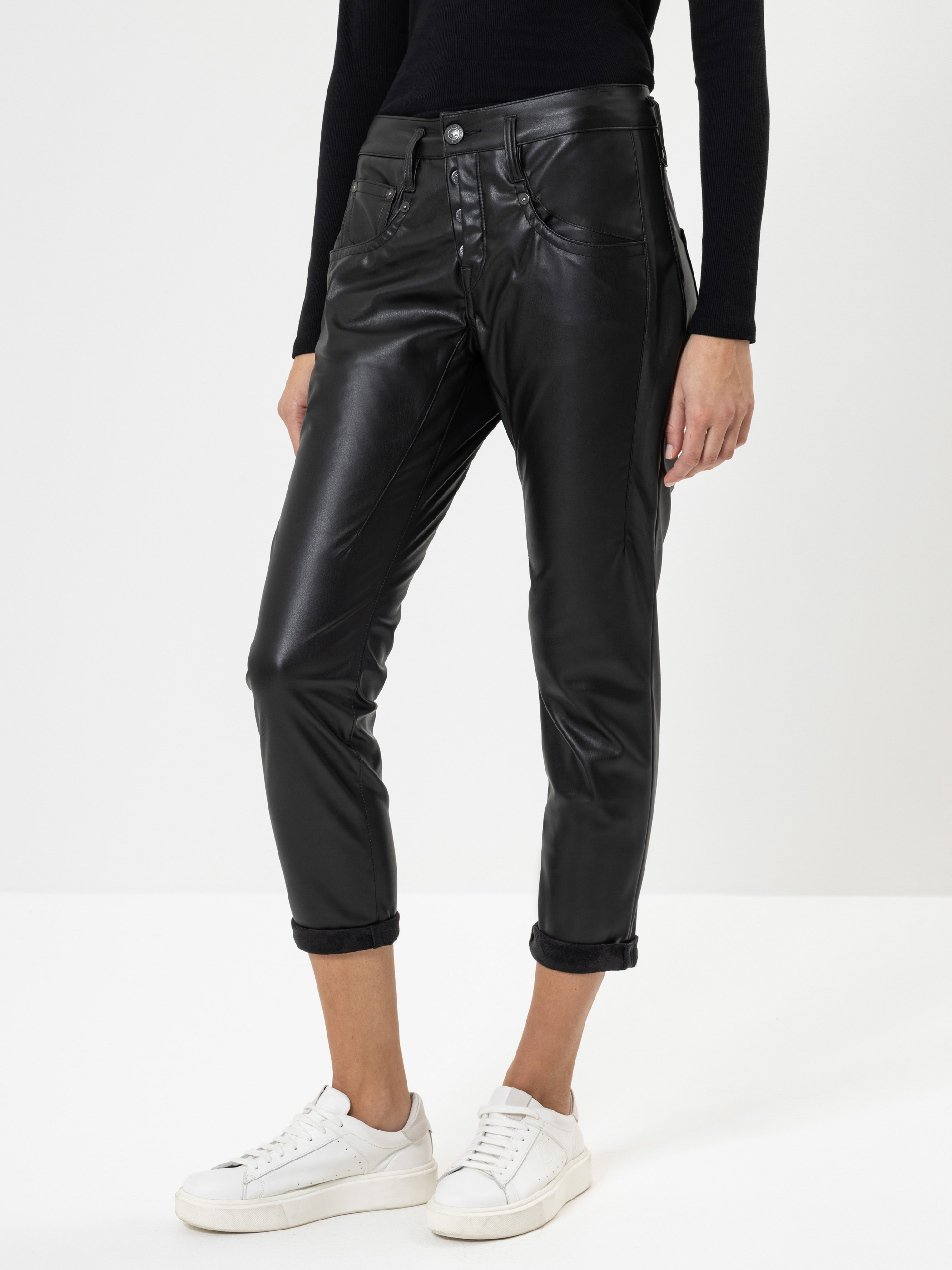 Eine Person, die eine schwarze Lederhose, ein schwarzes langärmeliges Oberteil und weiße Turnschuhe trägt, steht vor einem schlichten weißen Hintergrund. Die Hose ist leicht beschnitten und an den Knöcheln gerollt.