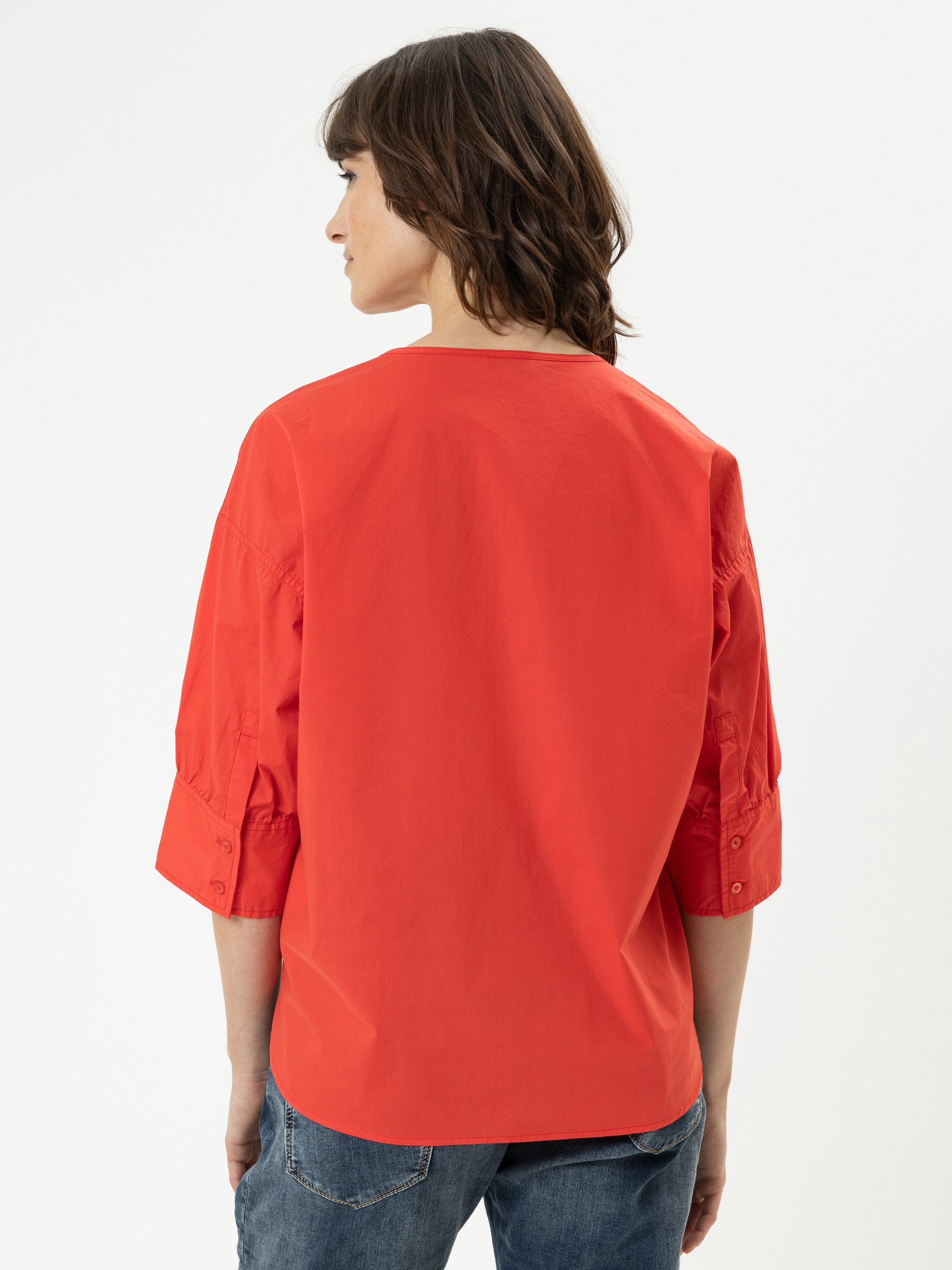 Eine Person mit schulterlangen braunen Haaren, die eine leuchtend rote, dreiviertelärmelige Bluse und blaue Jeans trägt, steht mit dem Rücken zur Kamera vor einem schlichten weißen Hintergrund.
