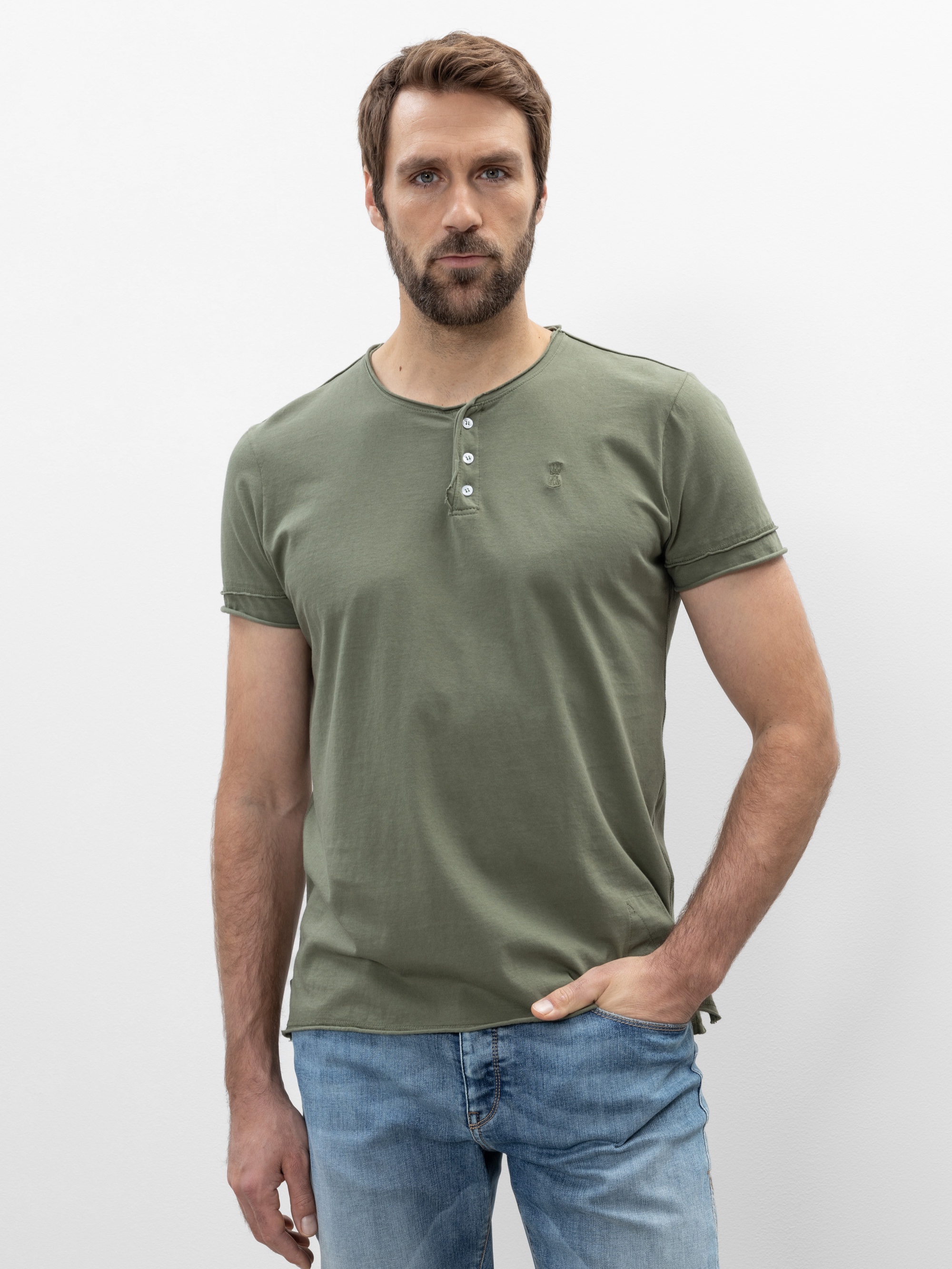 Ein Mann mit kurzen braunen Haaren und einem Bart steht vor einem schlichten weißen Hintergrund. Er trägt ein grünes kurzärmeliges Hemd und hellblaue Jeans und hat eine Hand in der Tasche.