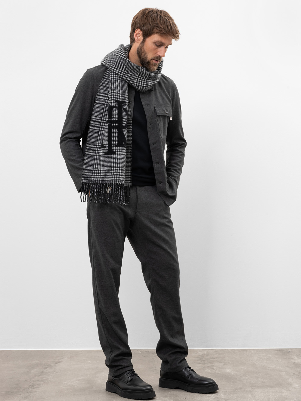 Ein Mann steht vor einer schlichten weißen Wand und trägt eine dunkelgraue Jacke, eine passende Hose, schwarze Schuhe und einen schwarz-weiß karierten Schal. Er blickt mit den Händen in den Taschen nach unten.