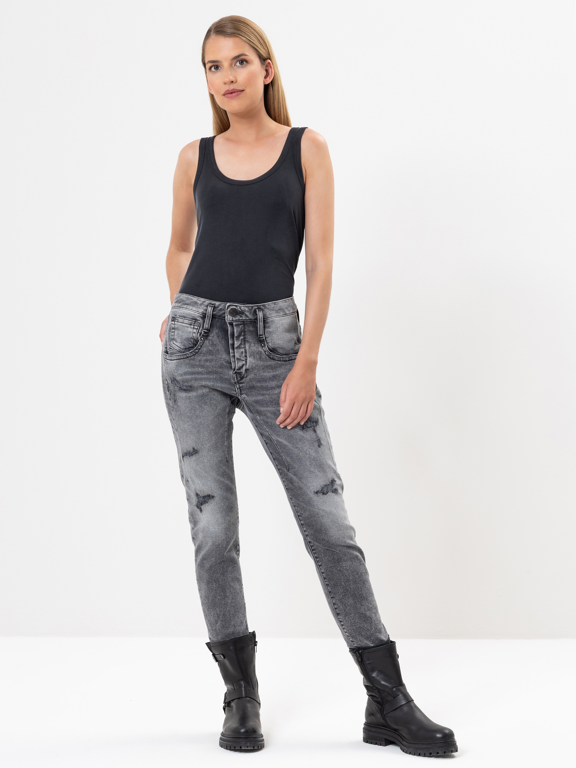 Eine Frau steht vor einem weißen Hintergrund und trägt ein schwarzes Tanktop, verwaschene graue Jeans mit abgenutzten Details und schwarze Kampfstiefel. Sie hat glattes, langes blondes Haar und die Hände in den Taschen.