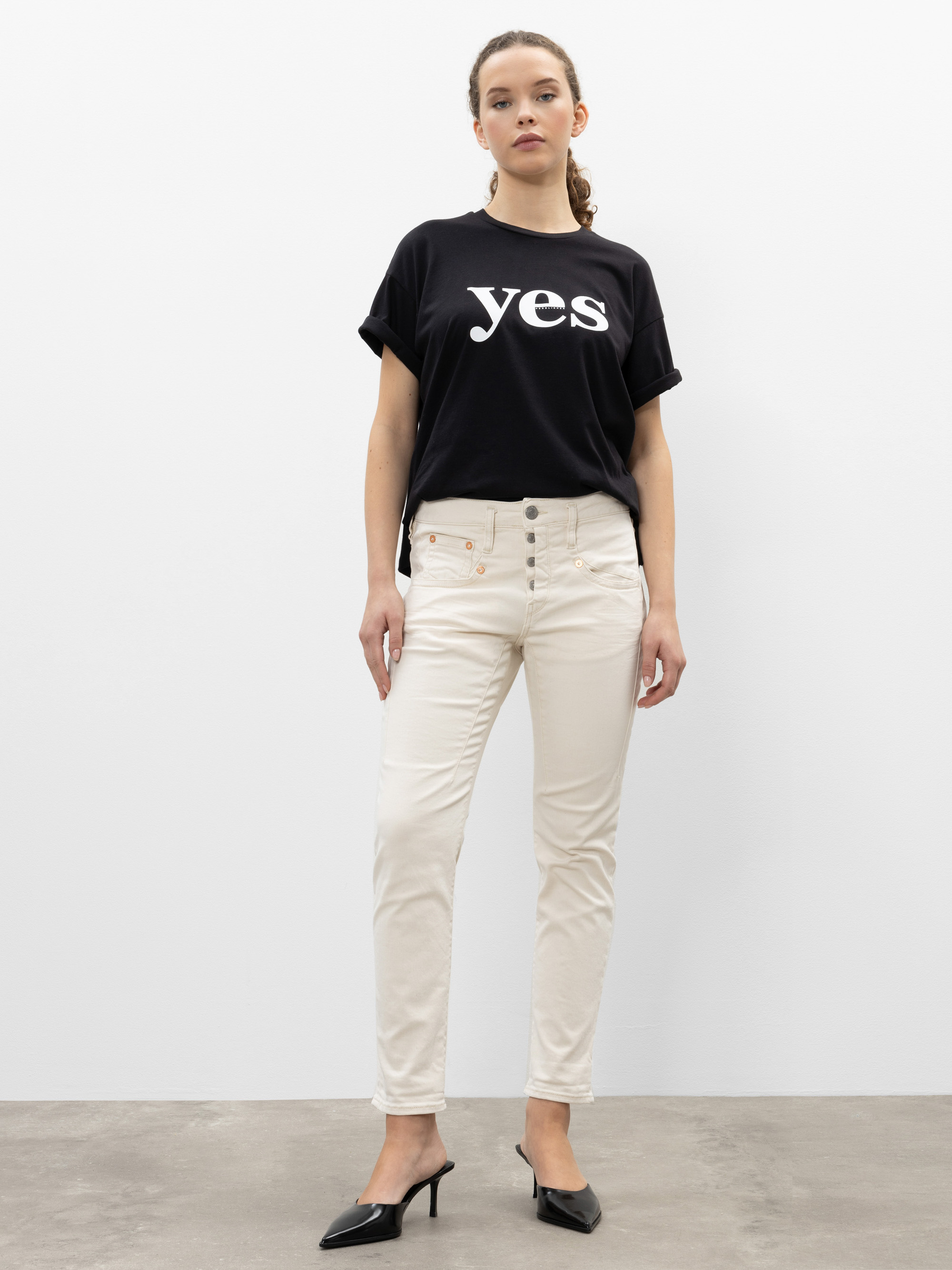 Eine Frau steht vor einer schlichten weißen Wand. Sie trägt ein schwarzes T-Shirt, auf dem in großen weißen Buchstaben "Ja" steht, eine cremefarbene Hose und schwarze Schuhe mit Absätzen. Sie hat ihr Haar zurückgebunden und steht mit neutraler Miene