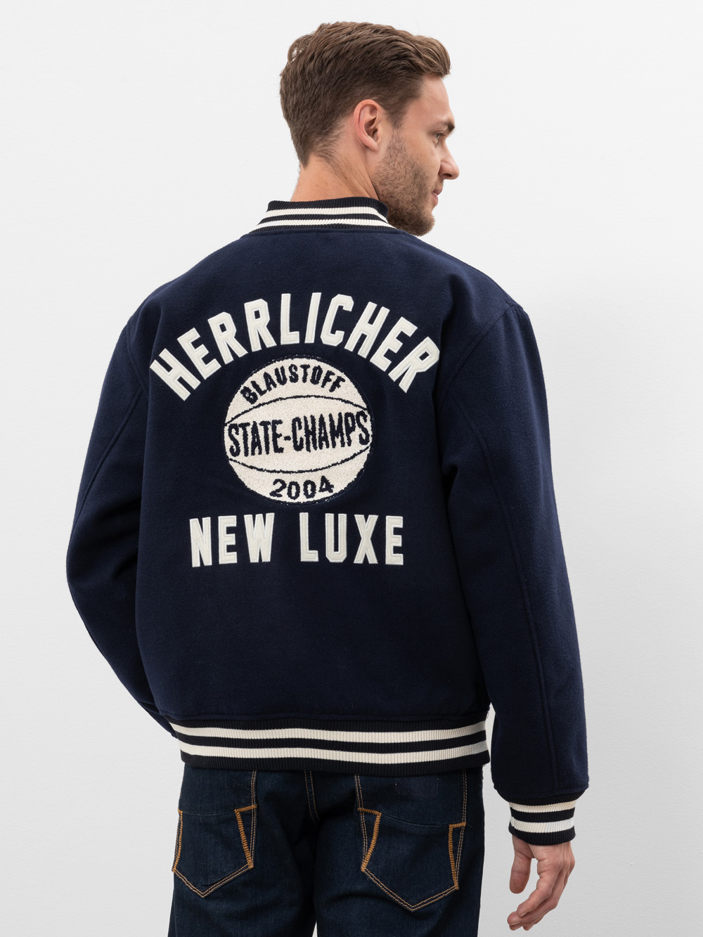 Ein Mann steht mit dem Gesicht zur Wand und trägt eine marineblaue Sportjacke mit weißem Saum. Auf der Rückseite der Jacke steht "HERRLICHER NEW LUXE" und ist ein kreisförmiger Aufnäher mit "BLAUSTOFF STATE-CHAMPS 2004" angebracht.