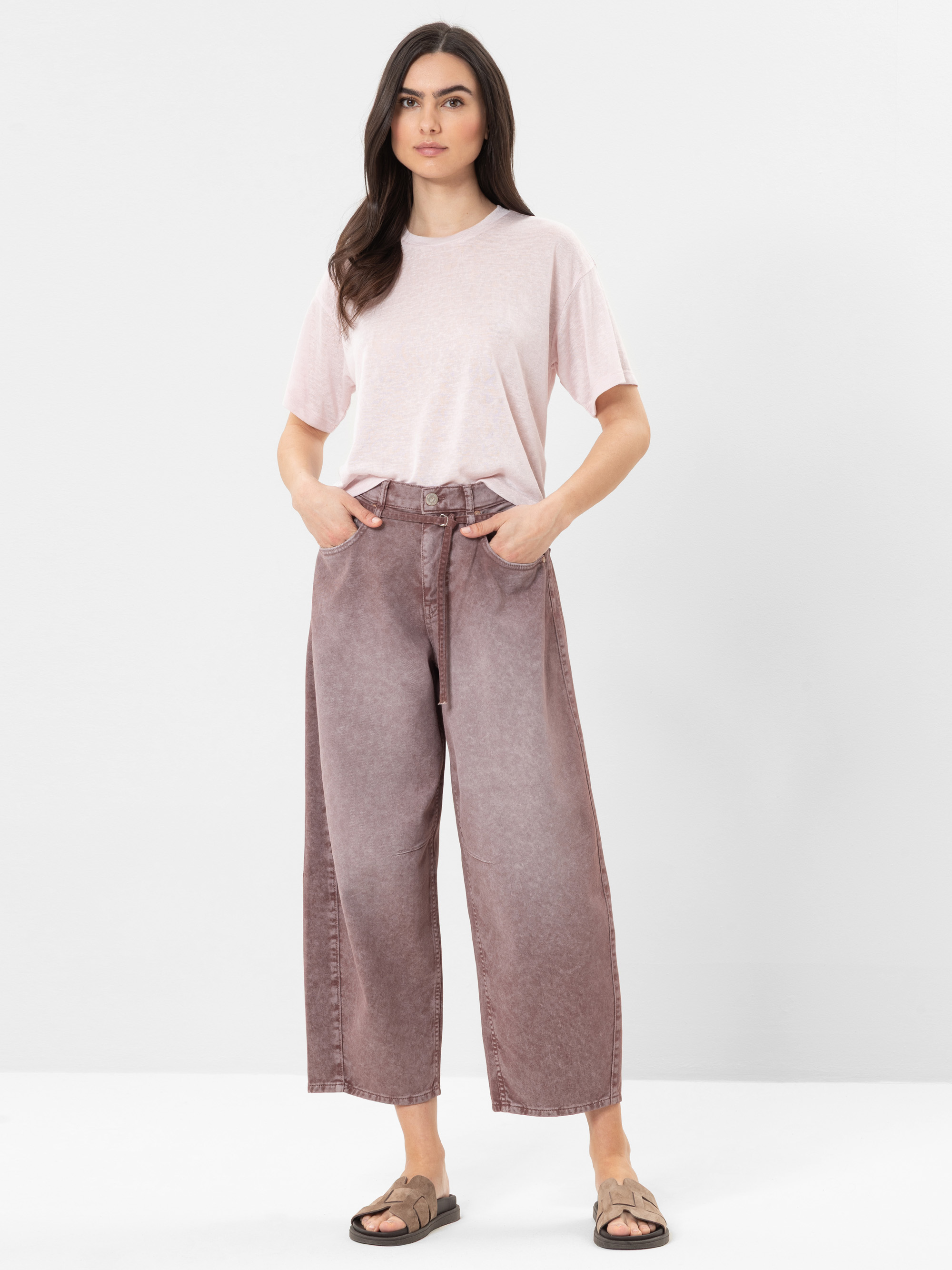 Eine Frau steht vor einem einfarbig weißen Hintergrund und trägt ein hellrosa T-Shirt, eine lilafarbene Jeans mit hoher Taille und weitem Bein sowie beigefarbene Sandalen. Sie hat lange dunkle Haare und ihre Hände stecken in den Jeanstaschen.