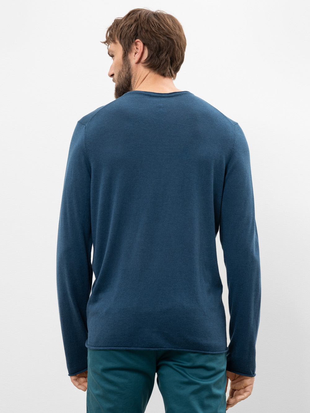 Ein Mann mit braunem Haar, der ein langärmeliges blaues Hemd und eine blaugrüne Hose trägt, steht mit dem Gesicht zur Kamera vor einem schlichten, hellen Hintergrund.