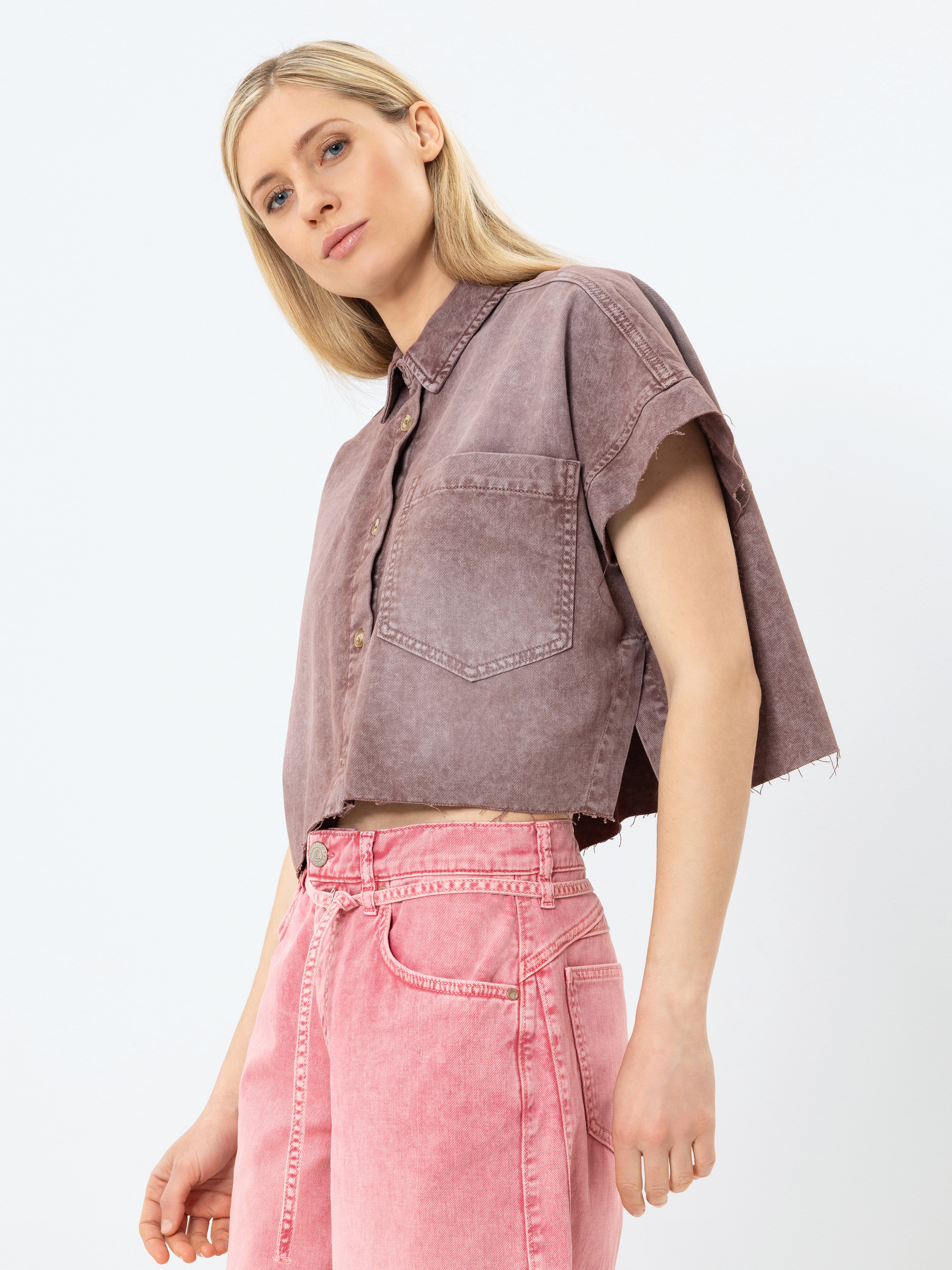 Eine Frau mit langen blonden Haaren trägt ein kurzärmeliges, lilafarbenes Jeanshemd und hochgekrempelte rosa Jeans. Sie steht vor einem schlichten weißen Hintergrund und blickt selbstbewusst in die Kamera.