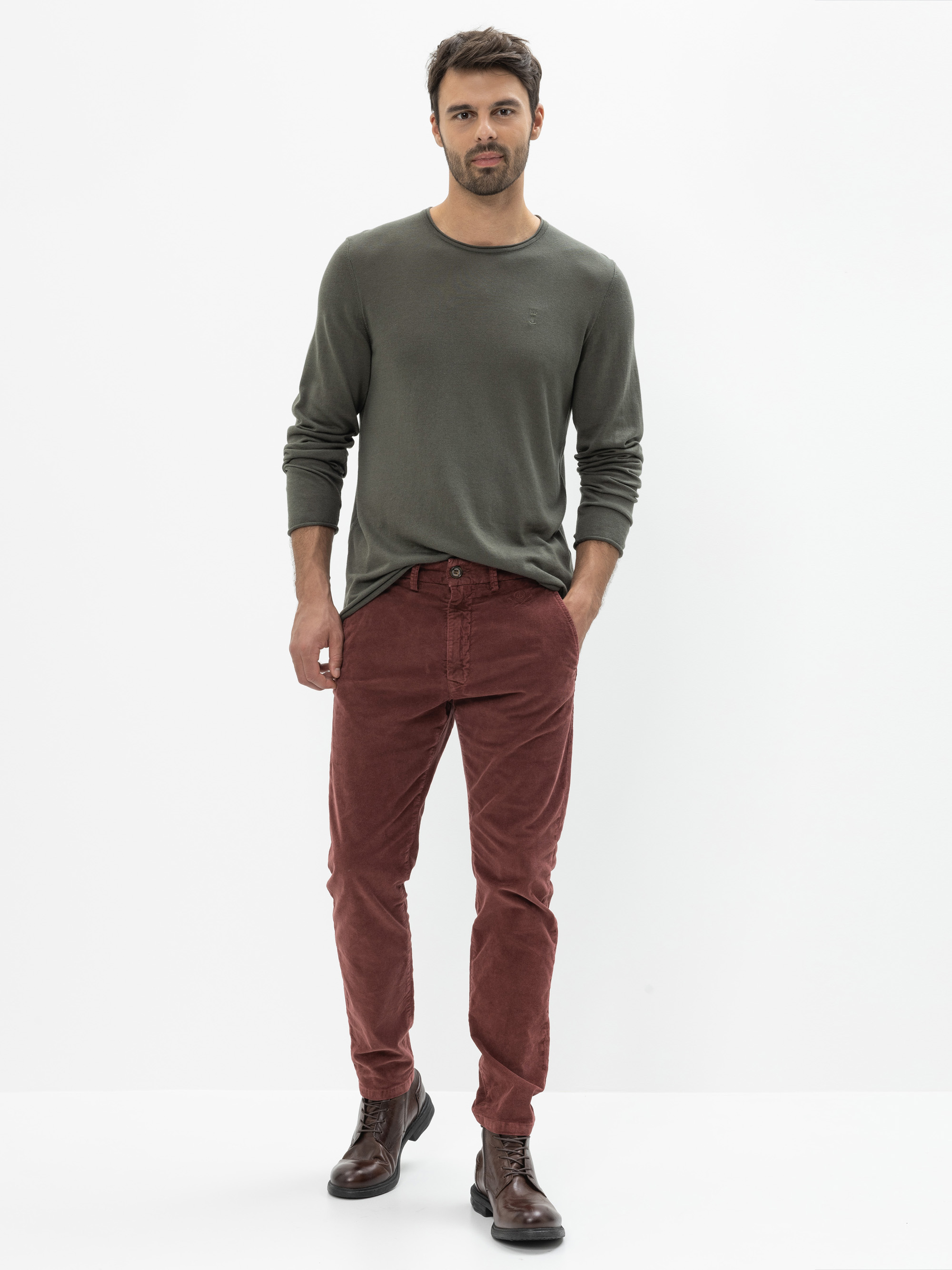 Ein Mann steht vor einem weißen Hintergrund und trägt ein langärmeliges olivgrünes Hemd, eine rostfarbene Cordhose und dunkelbraune Schnürstiefel. Er hat kurzes dunkles Haar und einen leichten Bart, eine Hand steckt in seiner Tasche.