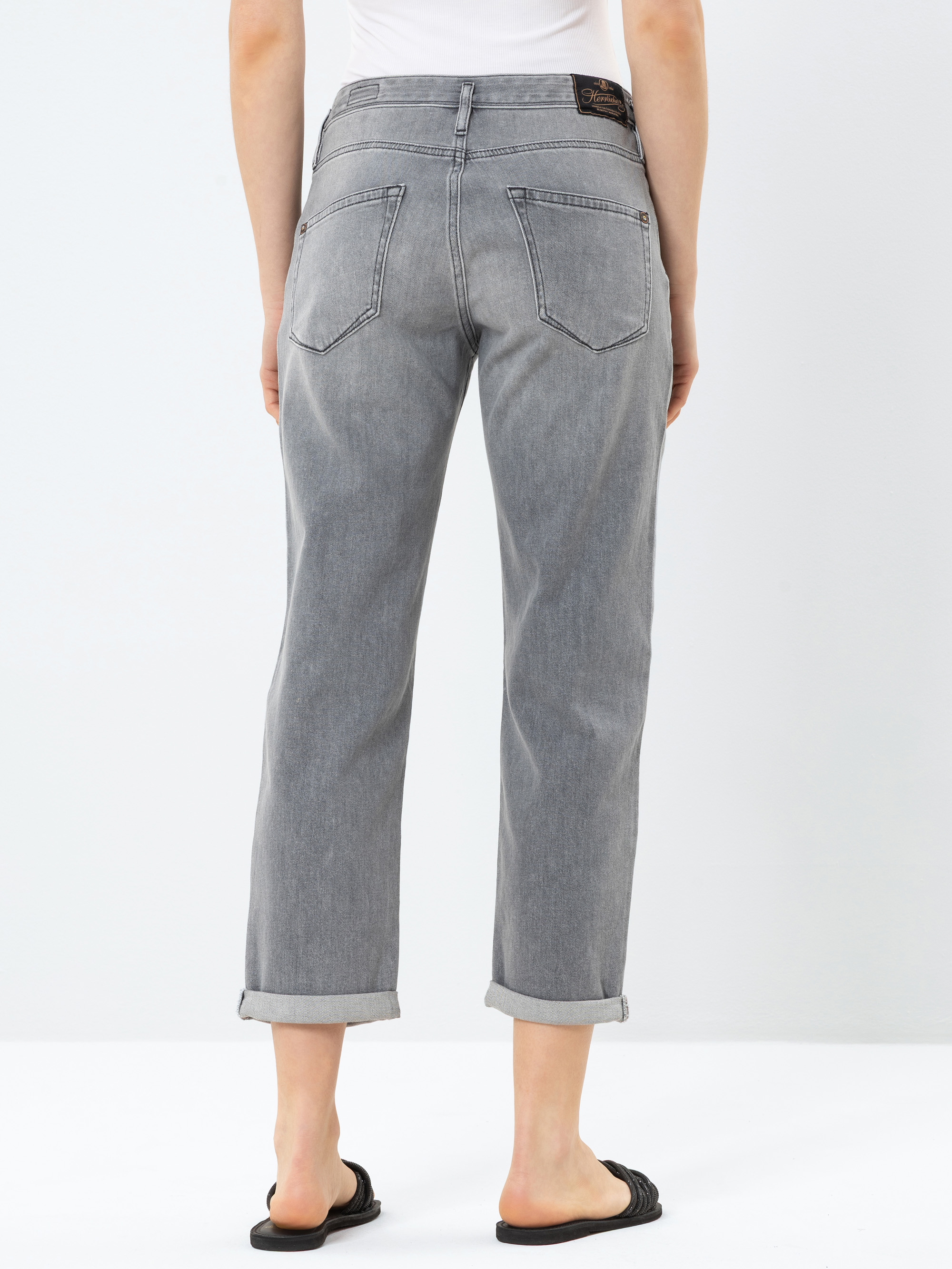 Eine Person, die hellgraue Jeans mit Bündchen und schwarze Sandalen trägt, steht mit dem Rücken zur Kamera vor einem schlichten weißen Hintergrund.