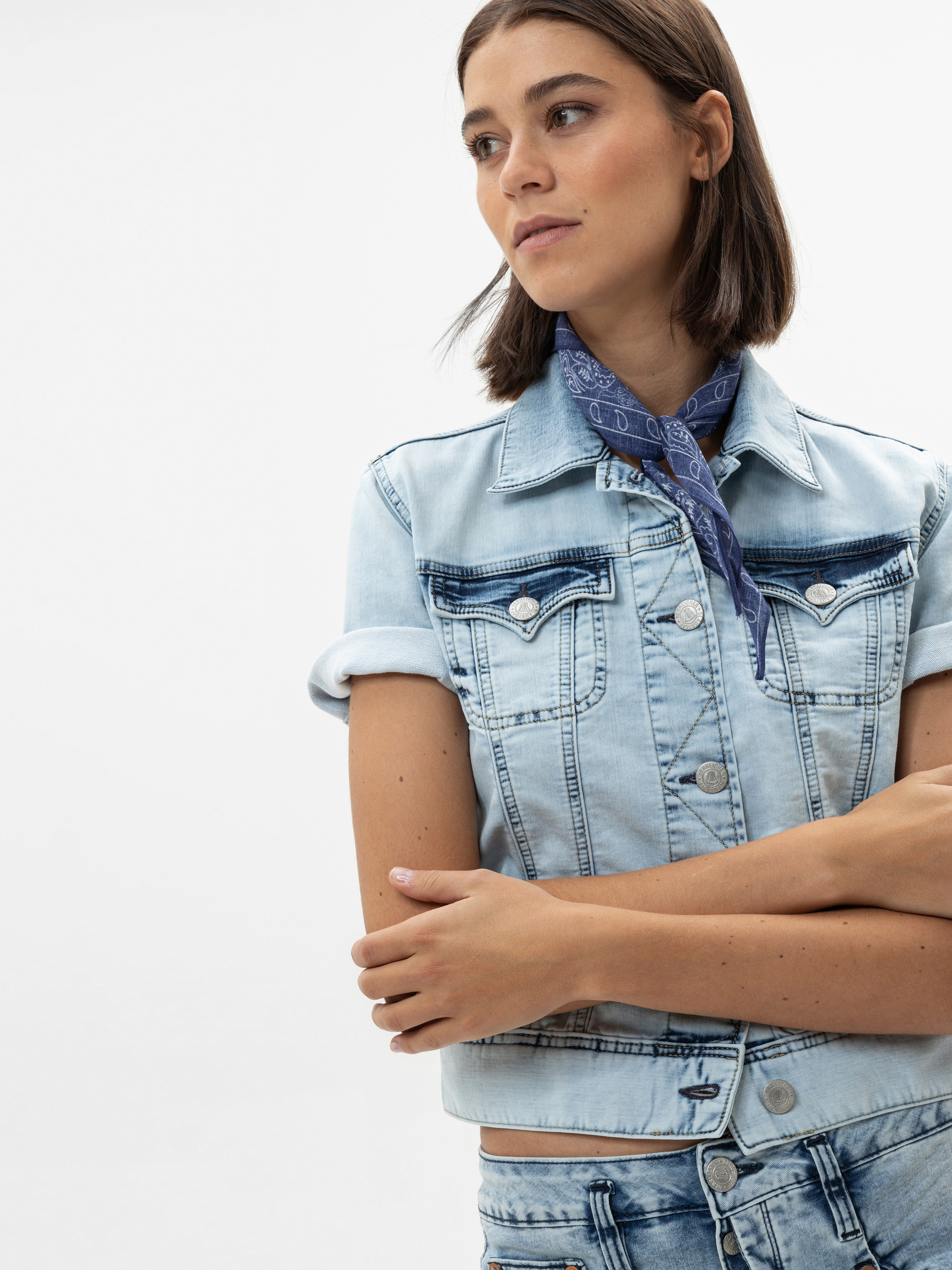 Eine Frau mit glattem braunem Haar trägt eine hellblaue Jeansjacke, passende Jeans und ein blaues Kopftuch um den Hals. Sie steht mit verschränkten Armen und blickt zur Seite vor einem schlichten weißen Hintergrund.