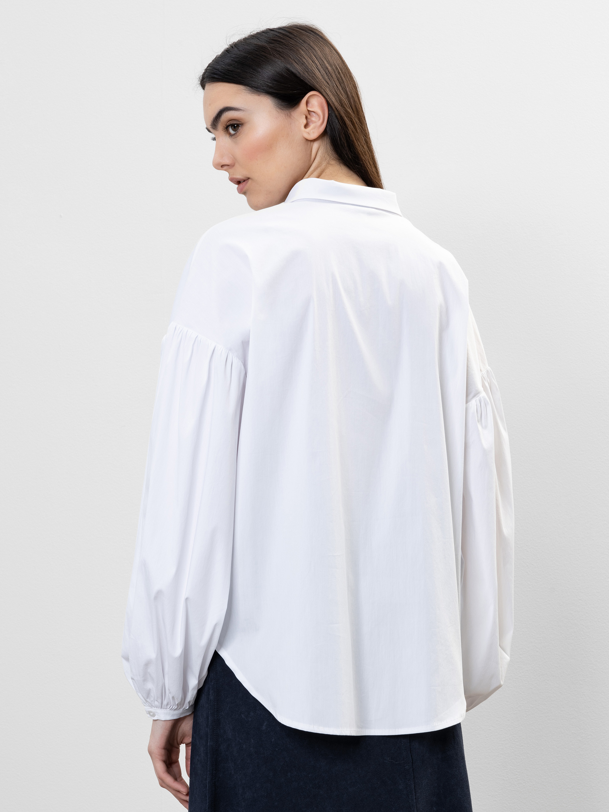 Eine Frau mit langen dunklen Haaren ist von hinten zu sehen. Sie trägt eine locker sitzende weiße Bluse mit Puffärmeln und Kragen, gepaart mit einem dunklen Rock vor einem schlichten hellen Hintergrund.
