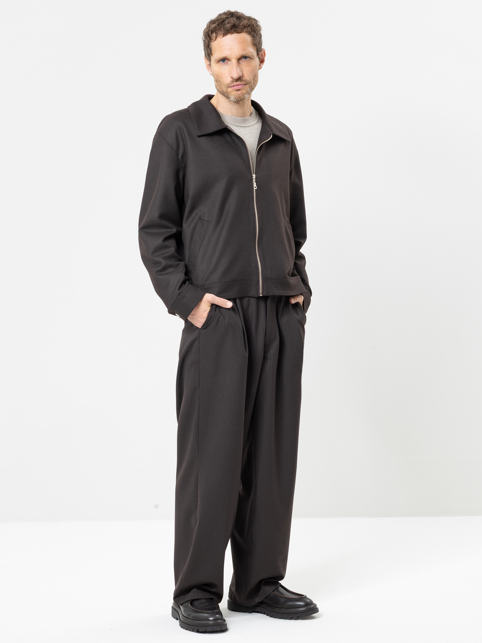 Vor einem weißen Hintergrund steht ein Mann mit den Händen in den Taschen; er trägt eine schwarze Jacke mit Reißverschluss, eine weite schwarze Hose, schwarze Schuhe und ein hellgraues Hemd.
