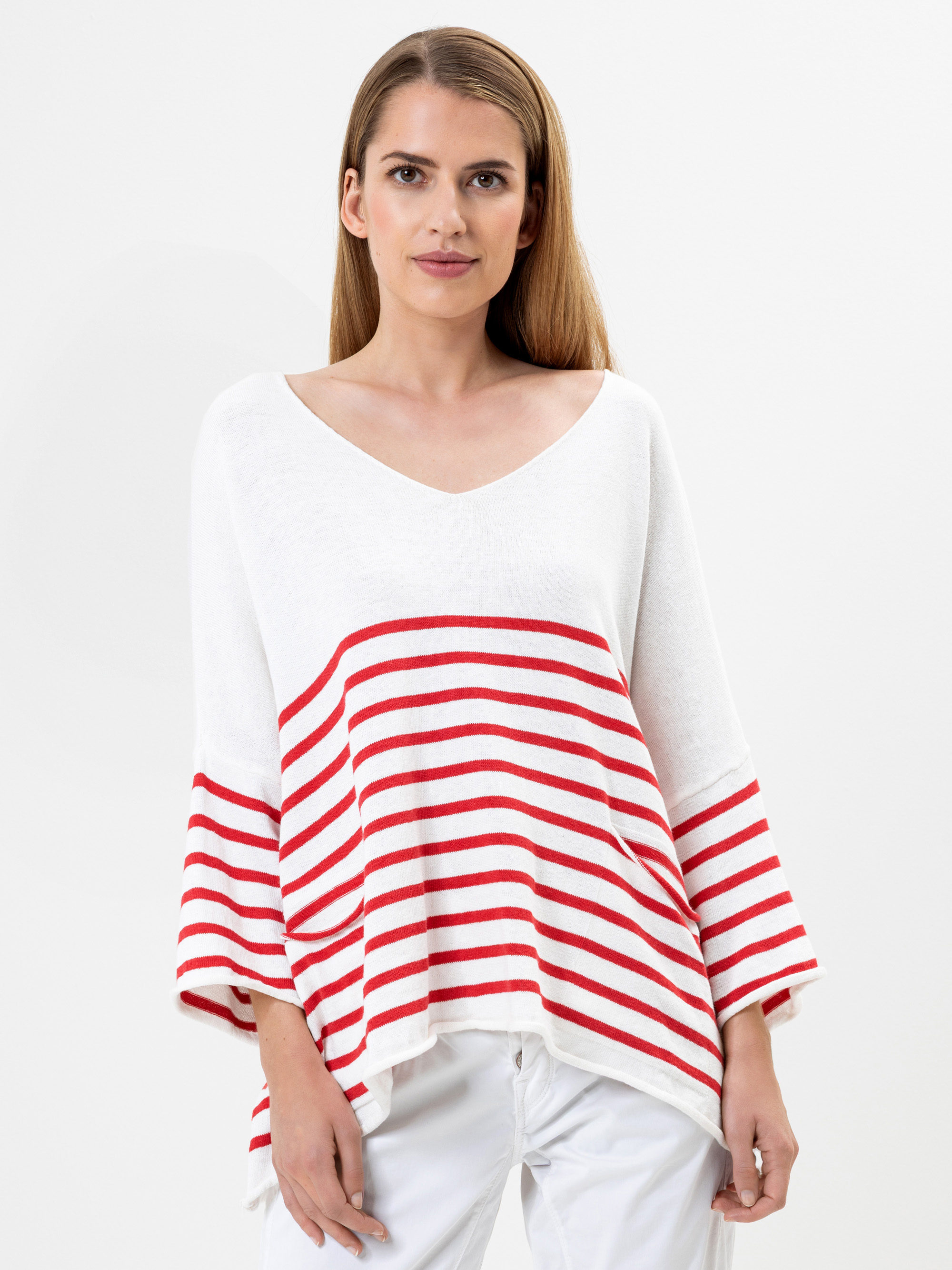 Eine Frau mit glattem, hellbraunem Haar trägt ein locker sitzendes weißes Oberteil mit roten Querstreifen und eine weiße Hose vor einem schlichten weißen Hintergrund.