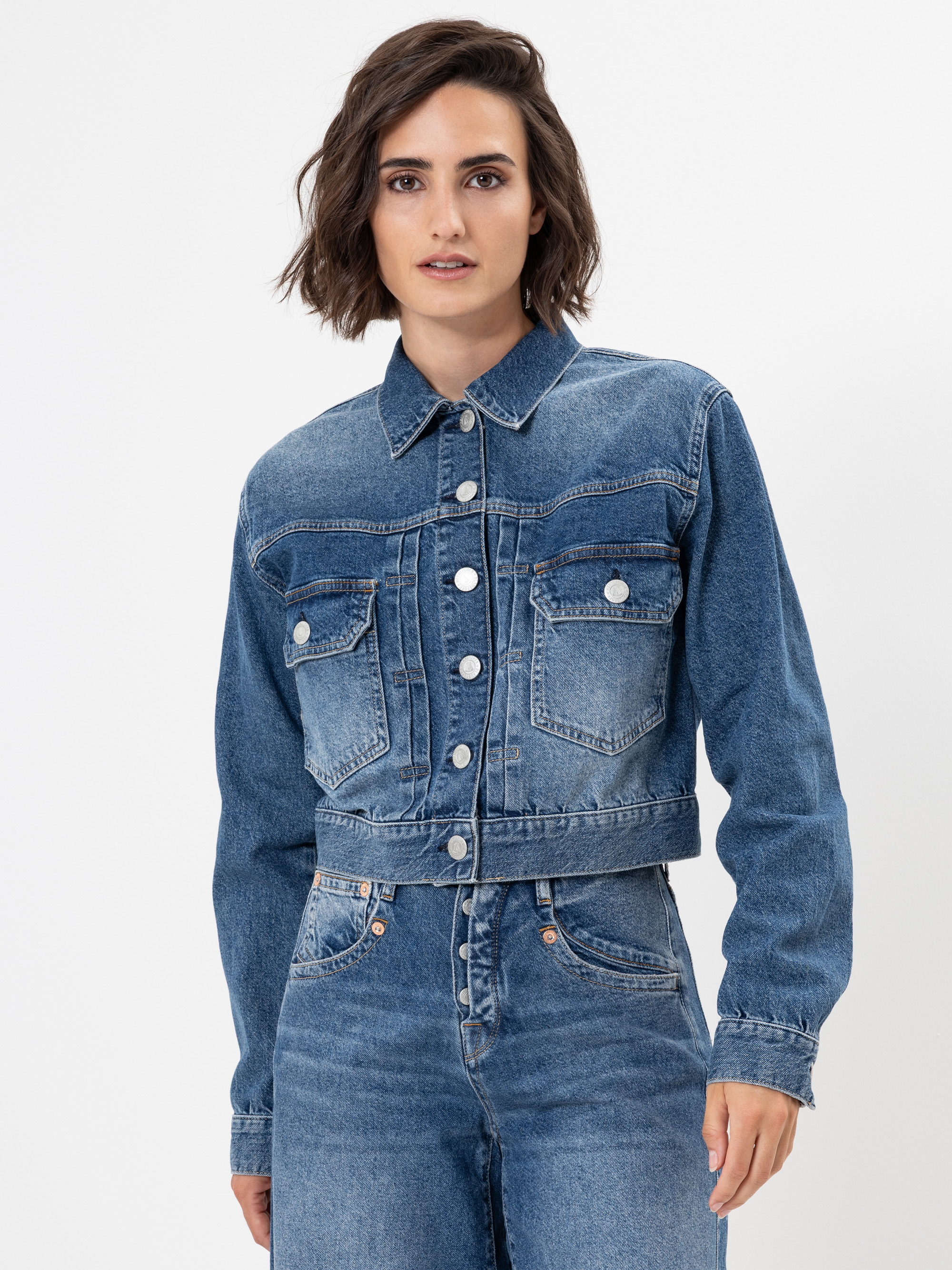 Eine Frau mit kurzen braunen Haaren trägt eine abgeschnittene blaue Jeansjacke mit Metallknöpfen und Vordertaschen, gepaart mit einer hochgeschnittenen Bluejeans, vor einem schlichten weißen Hintergrund.