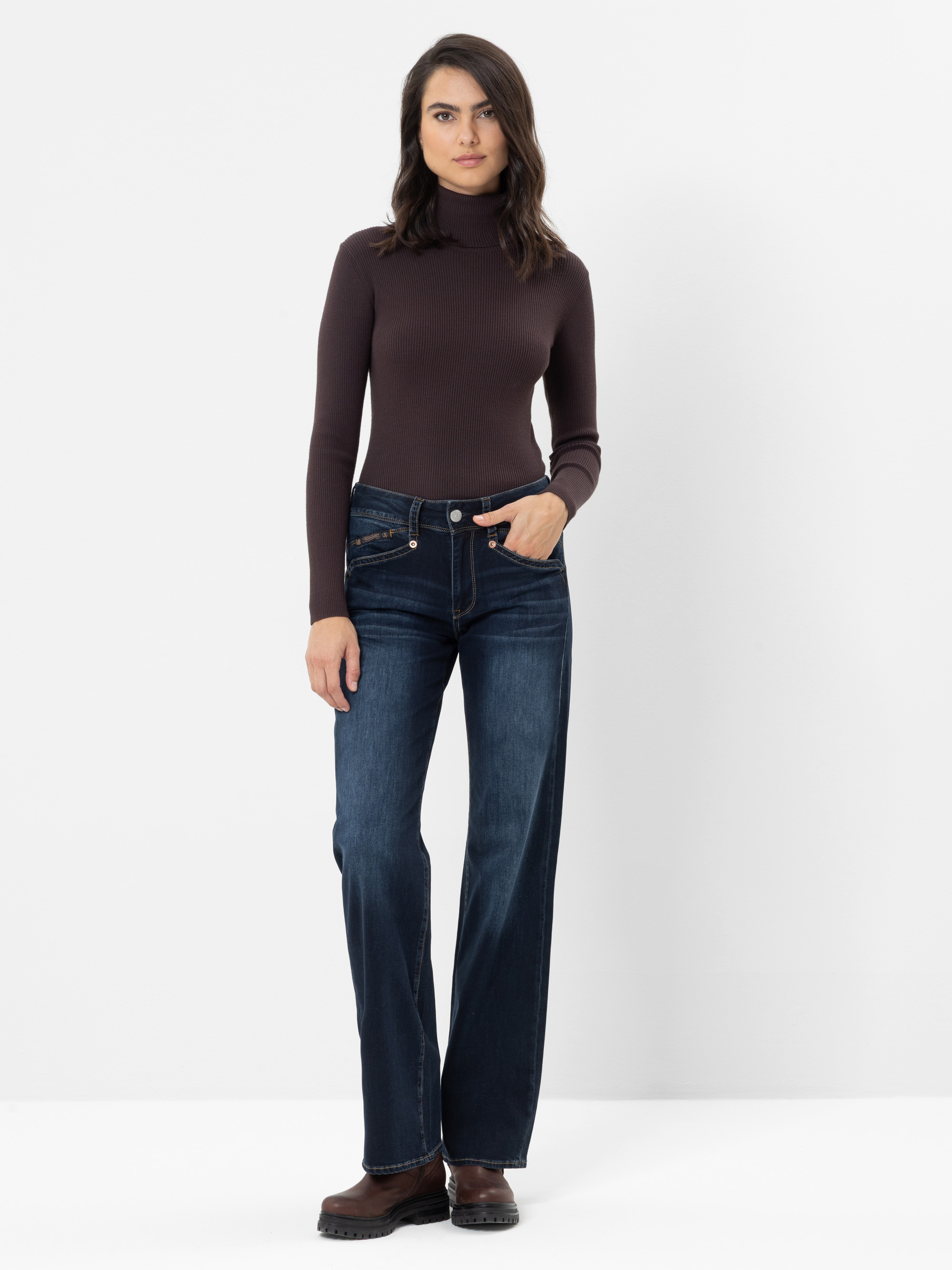 Eine Frau, die einen braunen Langarm-Rollkragenpullover, dunkelblaue Jeans mit weitem Bein und braune Schuhe trägt, steht vor einem schlichten weißen Hintergrund.