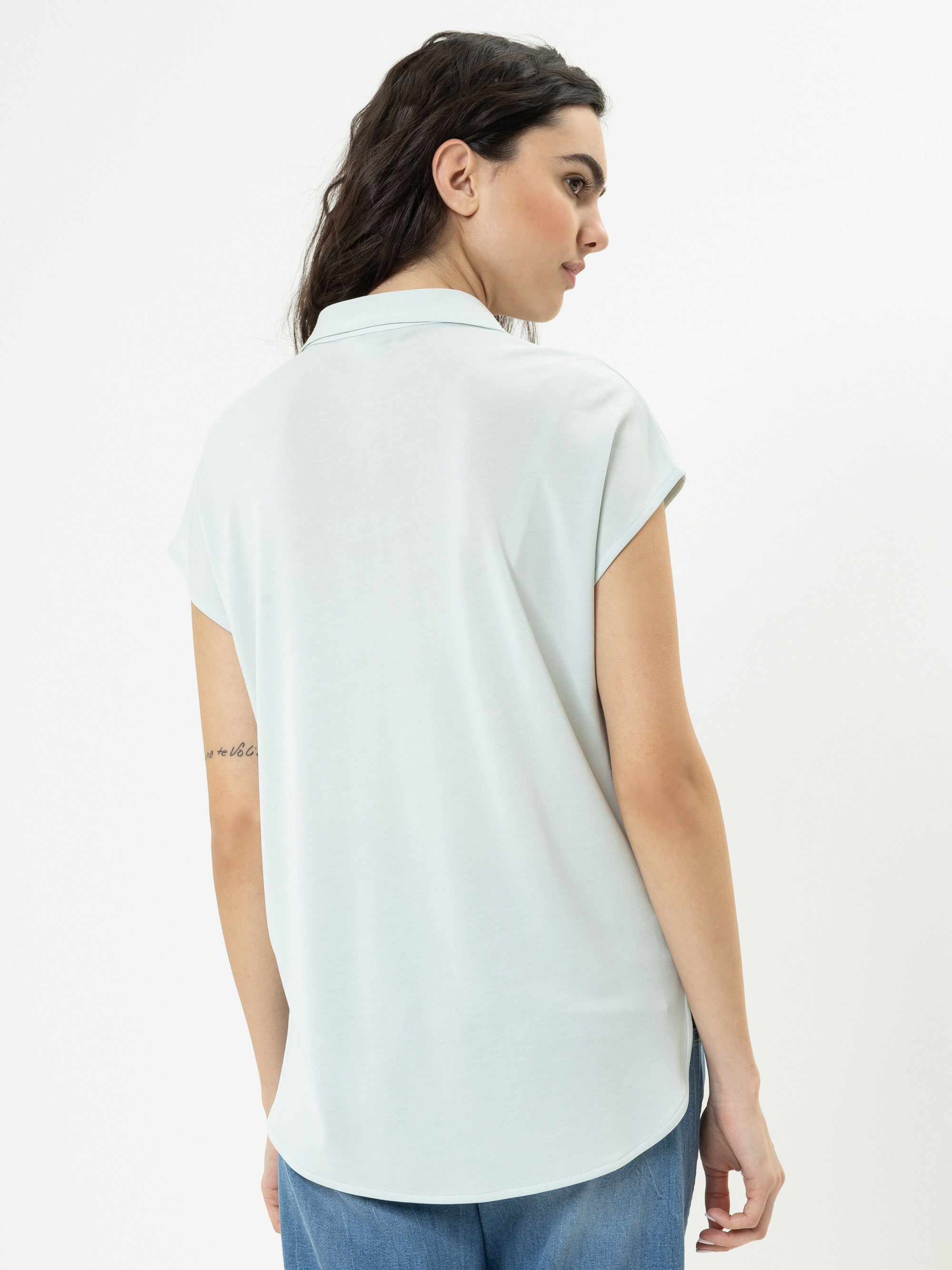 Eine Frau mit gewelltem dunklem Haar, bekleidet mit einem hellblauen kurzärmeligen Kragenhemd und blauen Jeans, steht von der Kamera weg. Auf ihrem rechten Oberarm ist eine Tätowierung zu sehen. Der Hintergrund ist einfarbig weiß.