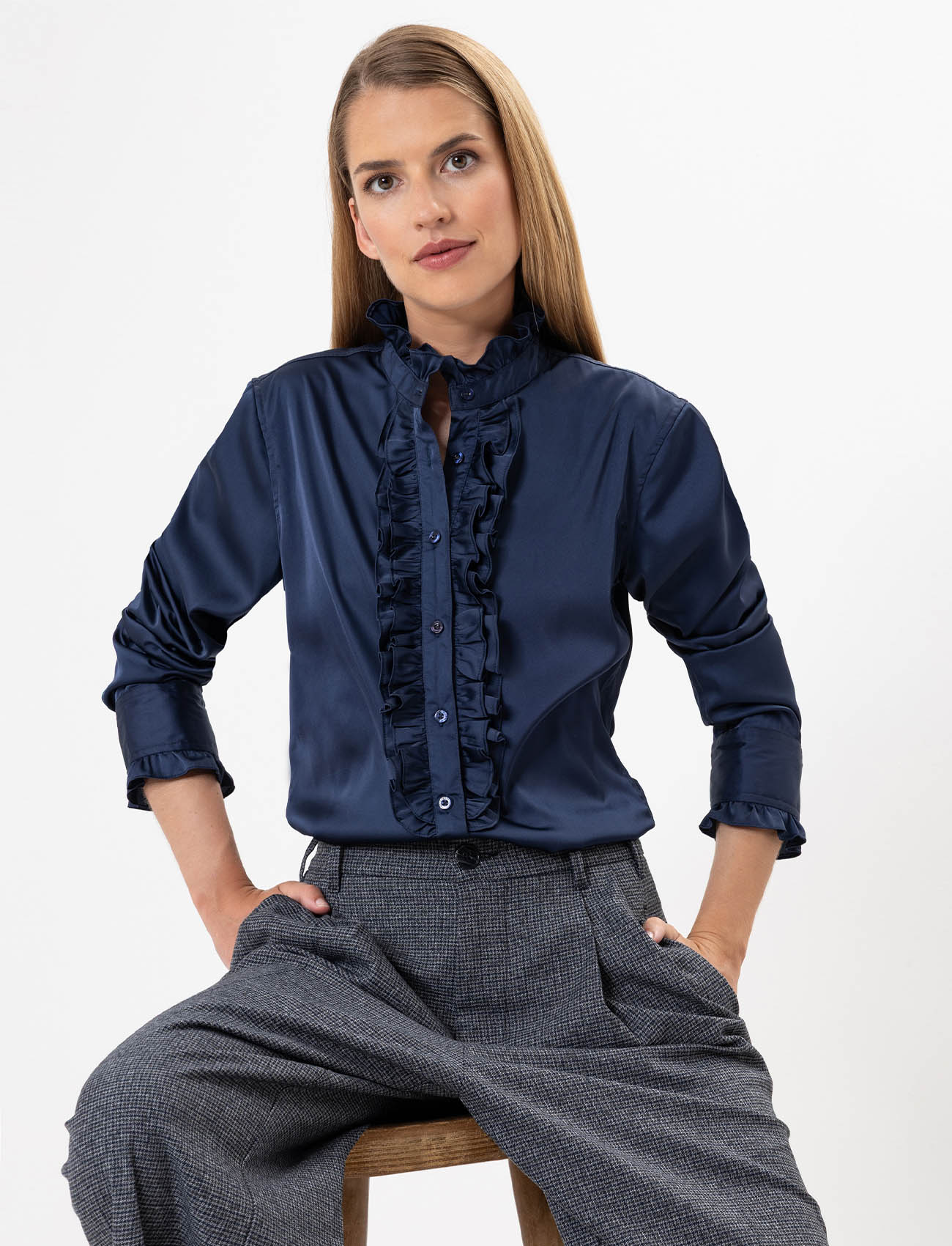 Eine Frau mit glattem, hellbraunem Haar sitzt auf einem Hocker und trägt eine marineblaue, langärmelige Bluse mit Rüschen und eine grau karierte Hose. Sie posiert mit den Händen in den Taschen vor einem schlichten weißen Hintergrund.