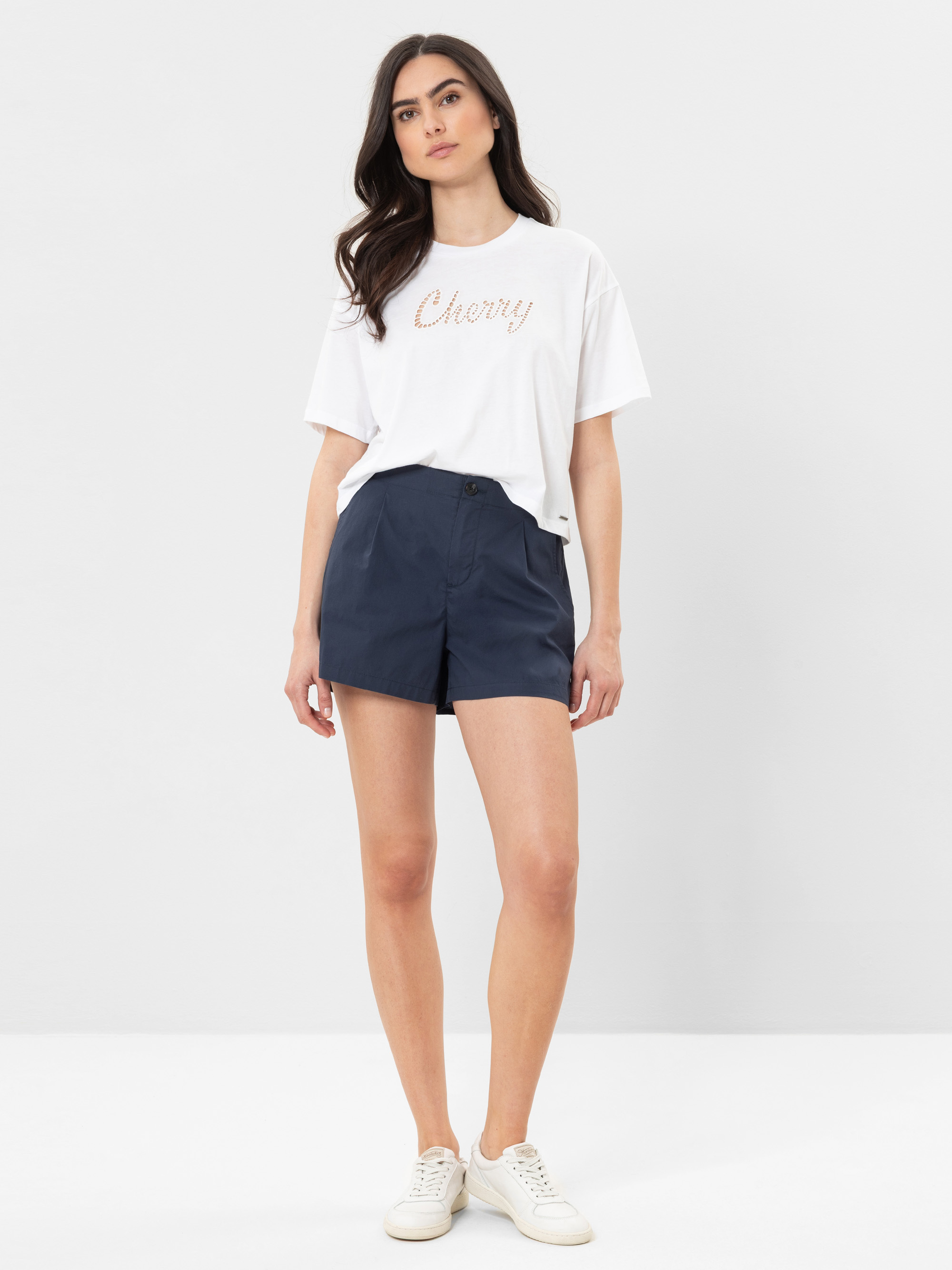 Eine Frau steht vor einem schlichten Hintergrund und trägt ein weißes T-Shirt mit der Aufschrift Cherry, marineblaue Shorts und weiße Turnschuhe.