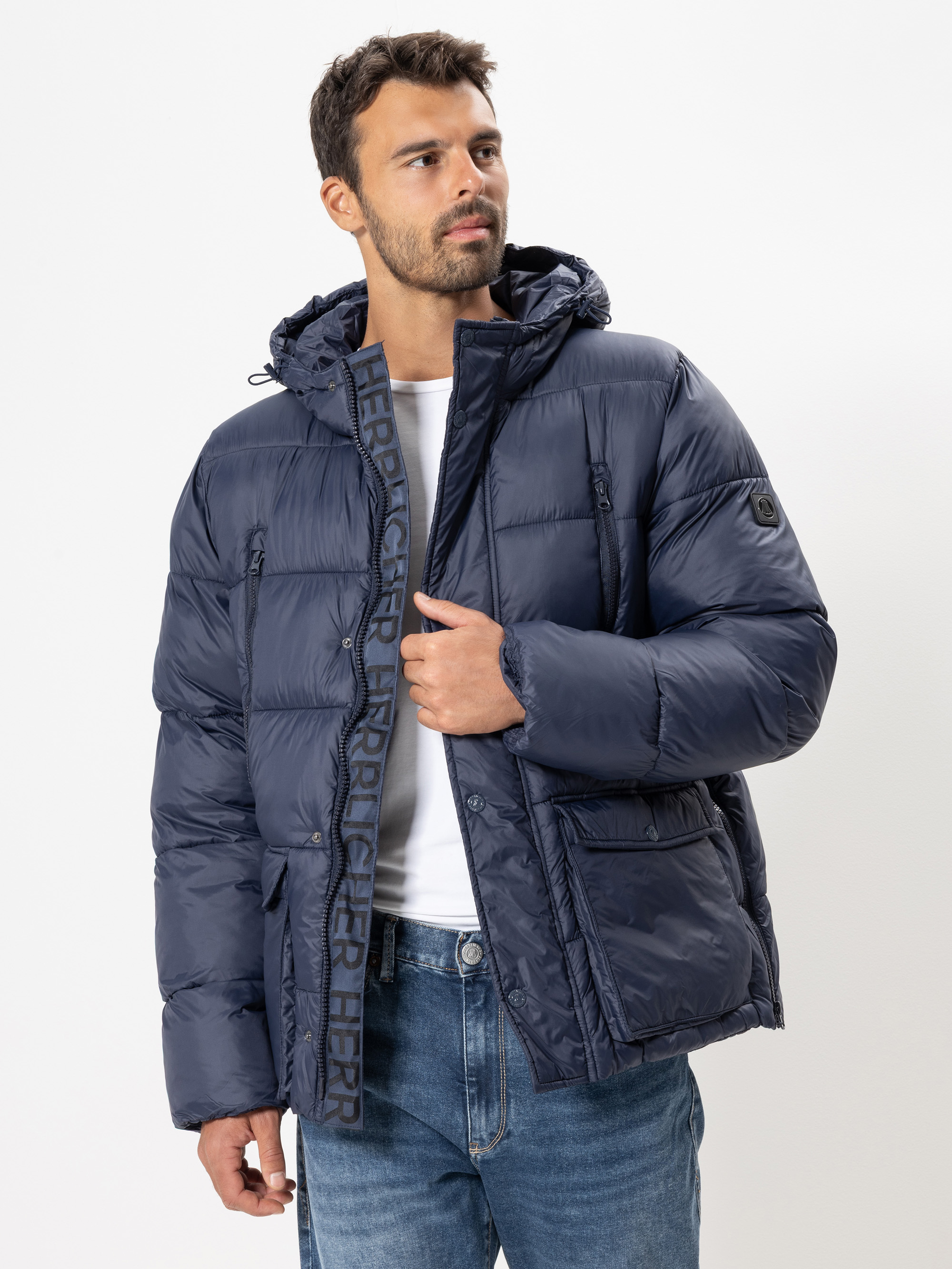 Ein Mann, der eine marineblaue Pufferjacke mit Kapuze und hellblaue Jeans trägt, steht vor einem schlichten weißen Hintergrund und blickt leicht zur Seite. Die Jacke hat einen sichtbaren Reißverschluss und Textdetails auf der Vorderseite.