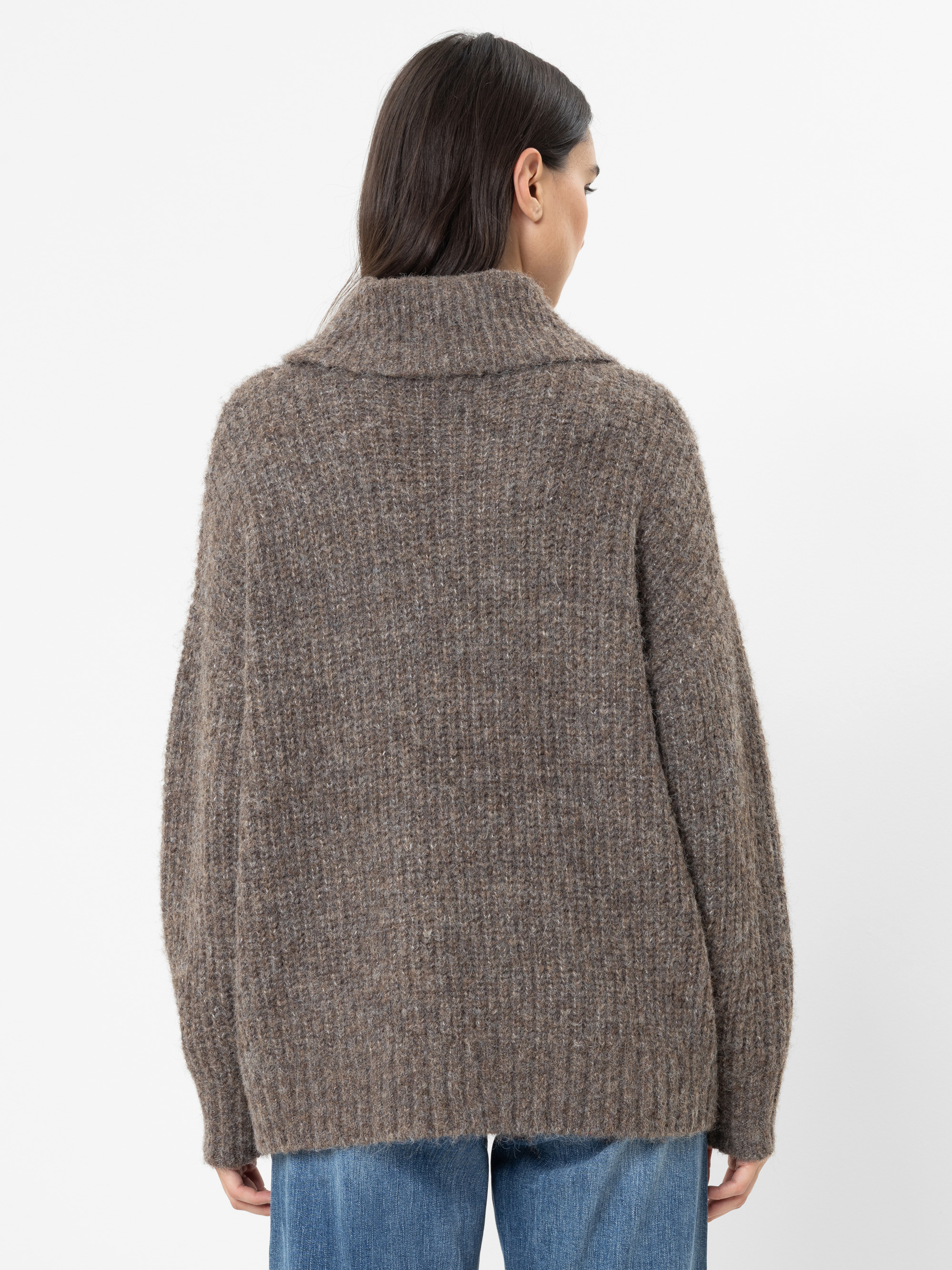 Eine Frau mit langen braunen Haaren steht mit dem Rücken zur Kamera und trägt einen lockeren, gerippten, braunen Strickpullover mit breitem Kragen. Außerdem trägt sie eine blaue Jeans. Der Hintergrund ist schlicht weiß.