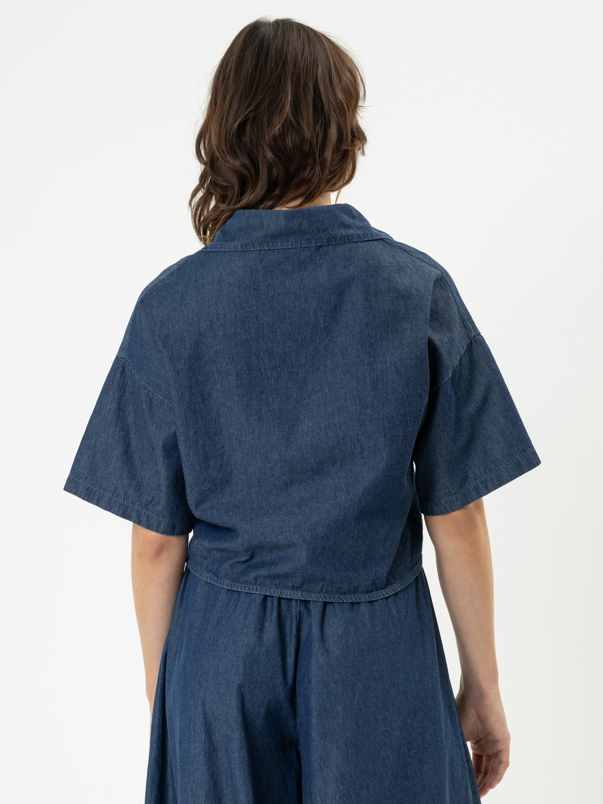 Eine Person mit gewelltem braunem Haar ist von hinten zu sehen, die ein kurzärmeliges, dunkles Jeansoberteil und eine dazu passende hochgeschnittene Hose vor einem schlichten weißen Hintergrund trägt.