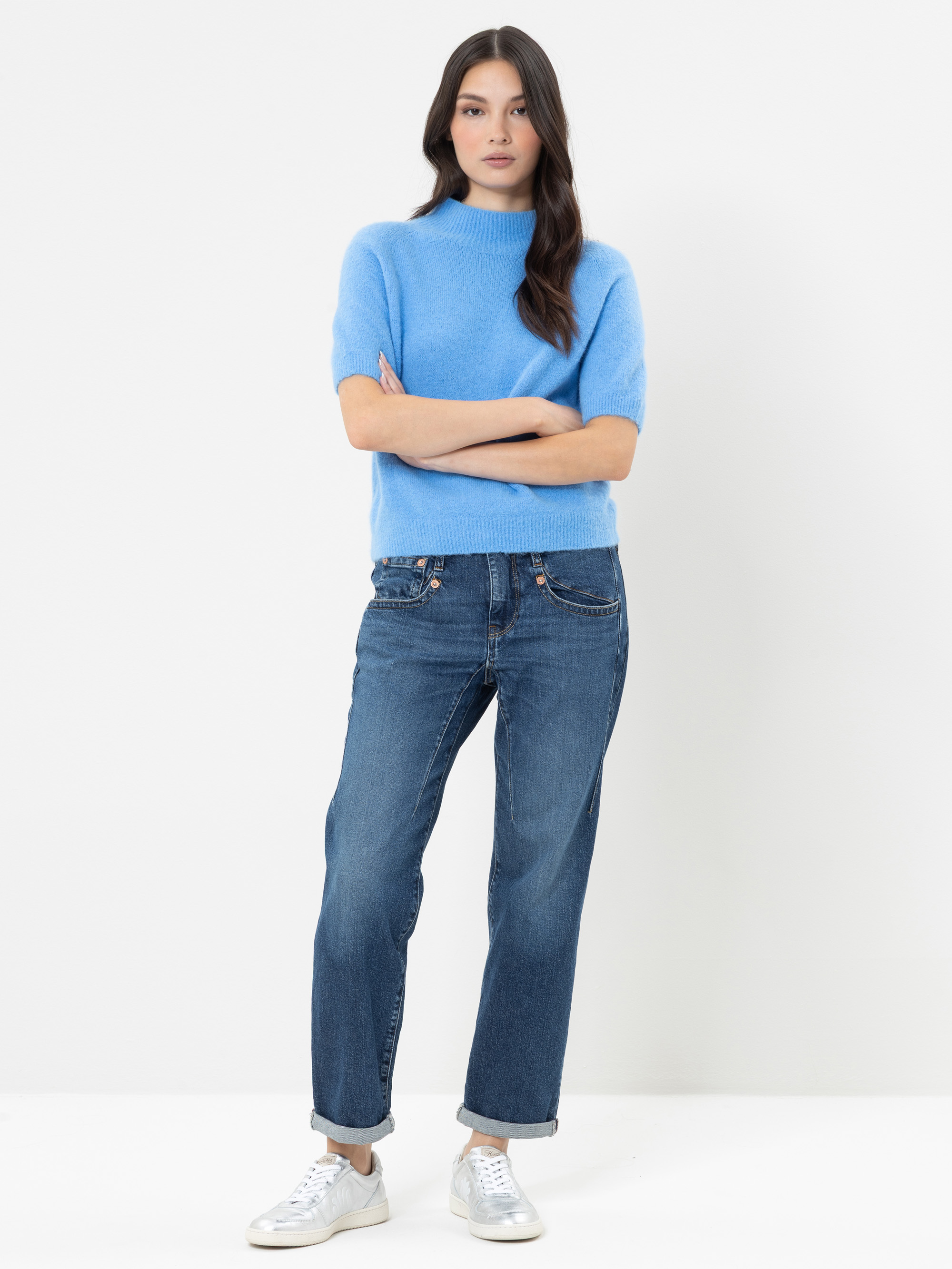 Eine Frau mit langen dunklen Haaren, die einen hellblauen, kurzärmeligen Pullover, eine blaue Jeans und weiße Turnschuhe trägt und mit verschränkten Armen vor einem schlichten weißen Hintergrund steht.